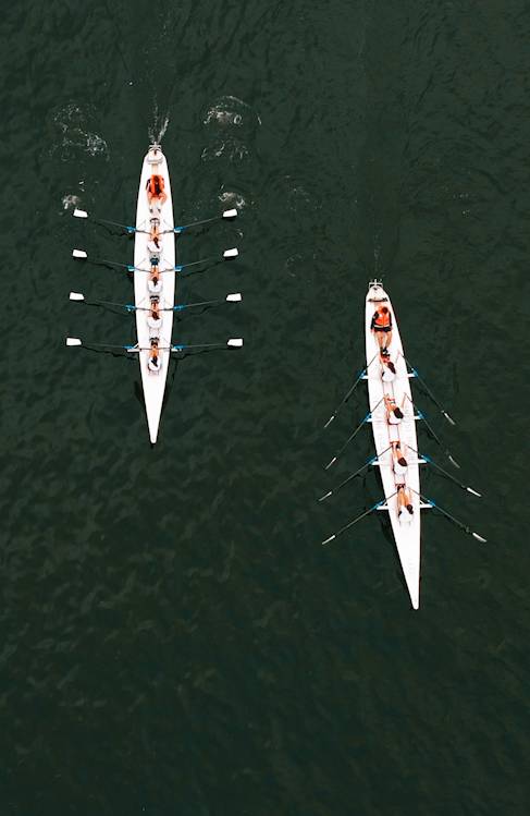 Two rowing teams paddle in parallel white boats on dark water, viewed from above near Rixos Tersane, each boat with eight rowers and one coxswain.