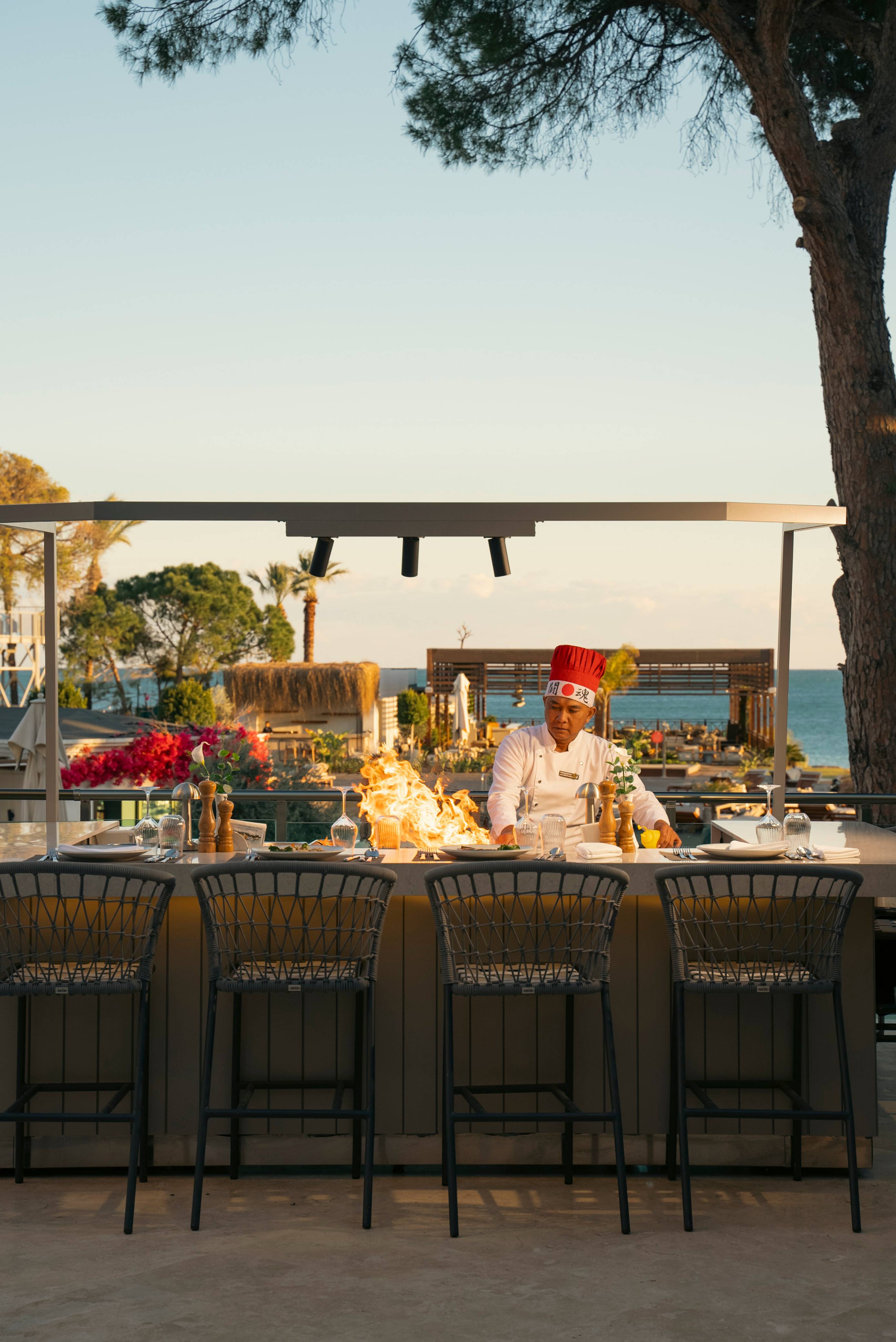 Chef performing a live teppanyaki show with flames at UMI Teppanyaki A La Carte Restaurant at Rixos Park Belek, surrounded by an outdoor dining setup with sea views.