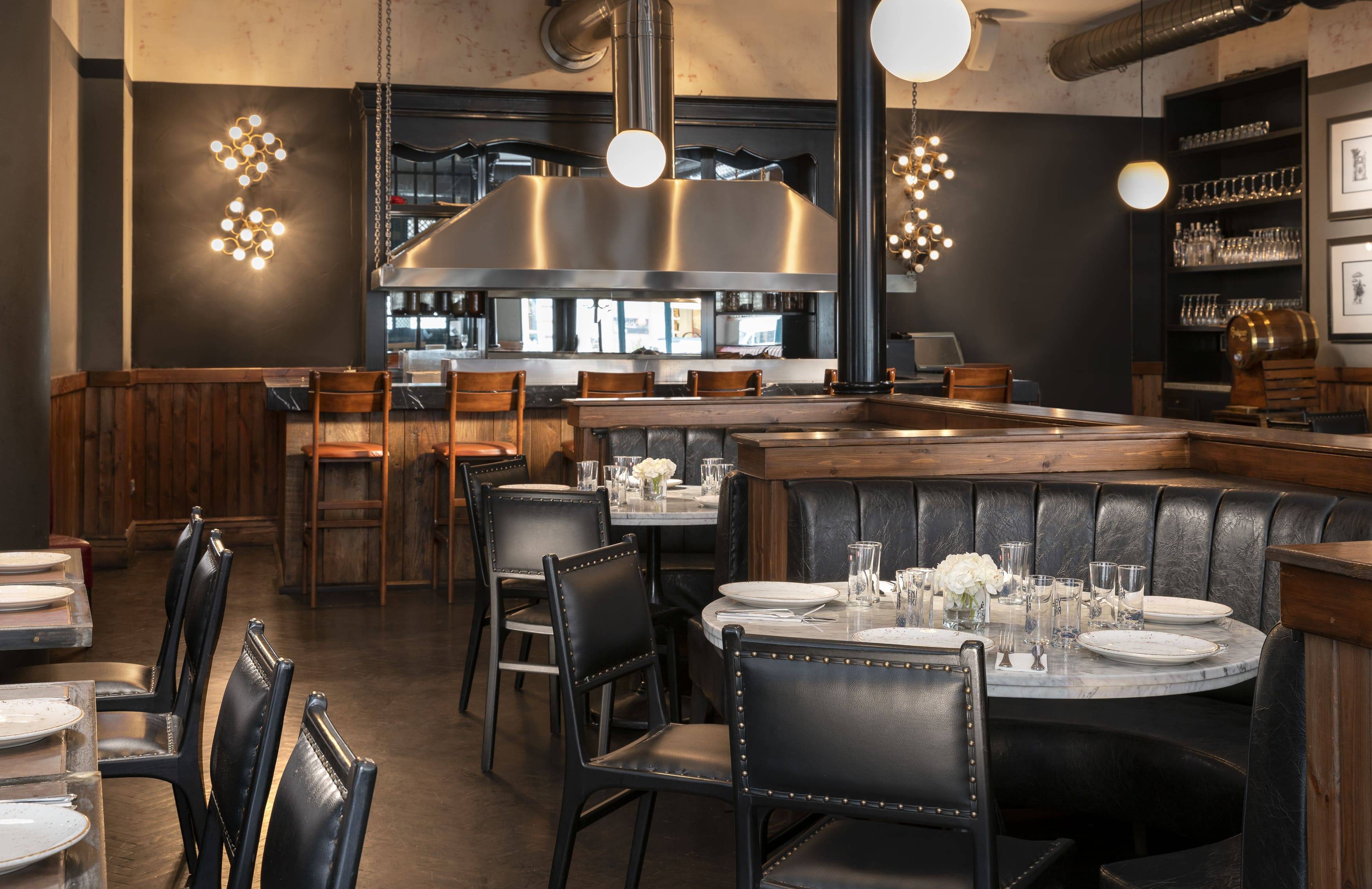 Snimka interijera restorana Kevok u Rixos Pera Istanbul, koji prikazuje topao, moderan industrijski dizajn. Blagovaonica ima polukružne crne kožne sjedalice i mramorne stolove postavljene za objedovanje, okružene crnim kožnim stolicama. U pozadini je vidljiv barski prostor s drvenim barskim stolicama, kojim dominira velika napa od nehrđajućeg čelika. Zidovi su tamno sivi i djelomično prekriveni drvenim panelima, osvijetljeni elegantnim, mesinganim zidnim svjetiljkama s više žarulja i jednostavnom kuglastom olovkom.