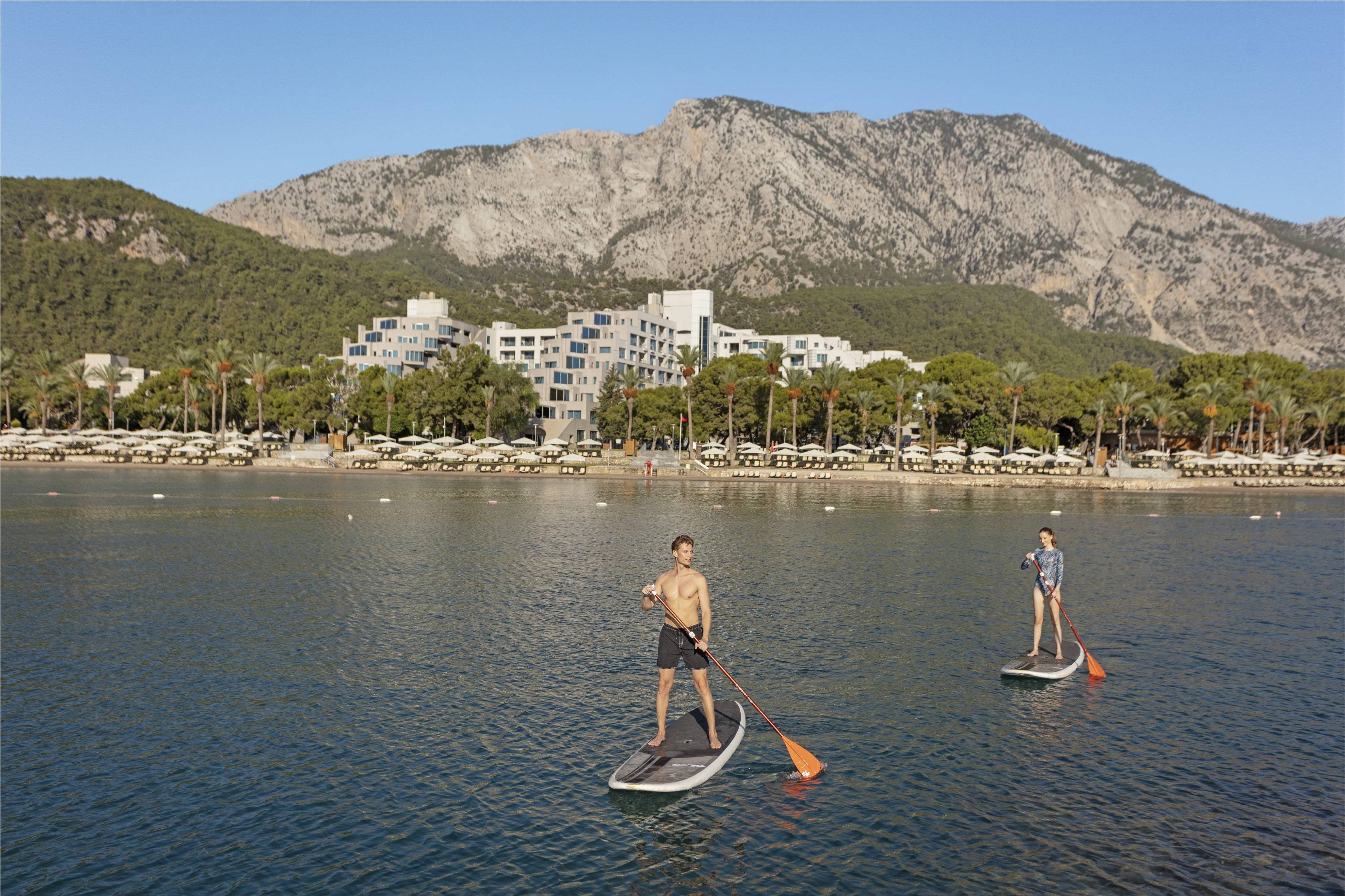 Two people paddleboarding on the sea in front of the Rixos Sungate hotel complex, which is backed by a large, forested mountain