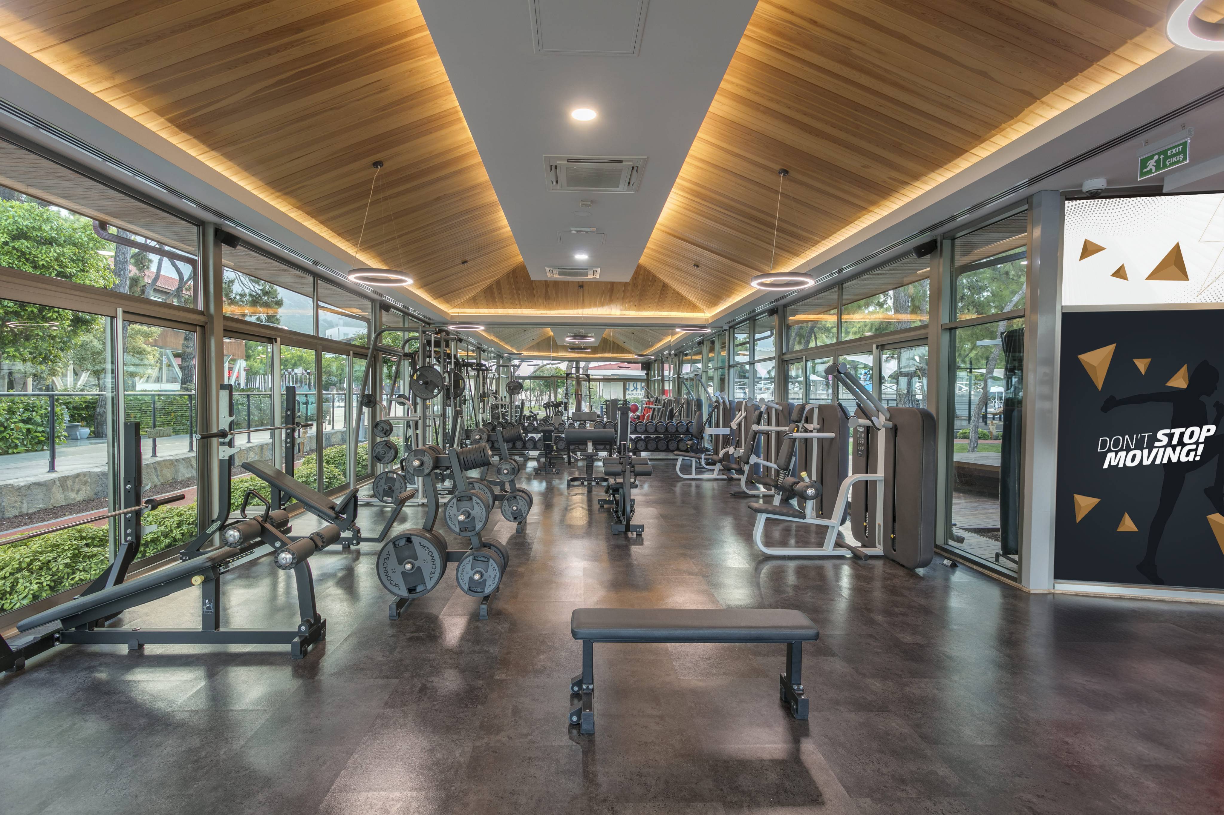 A modern, well-equipped gymnasium with a dark floor, large windows, and a slanted wood-paneled ceiling, showing various weight and cardio machines.