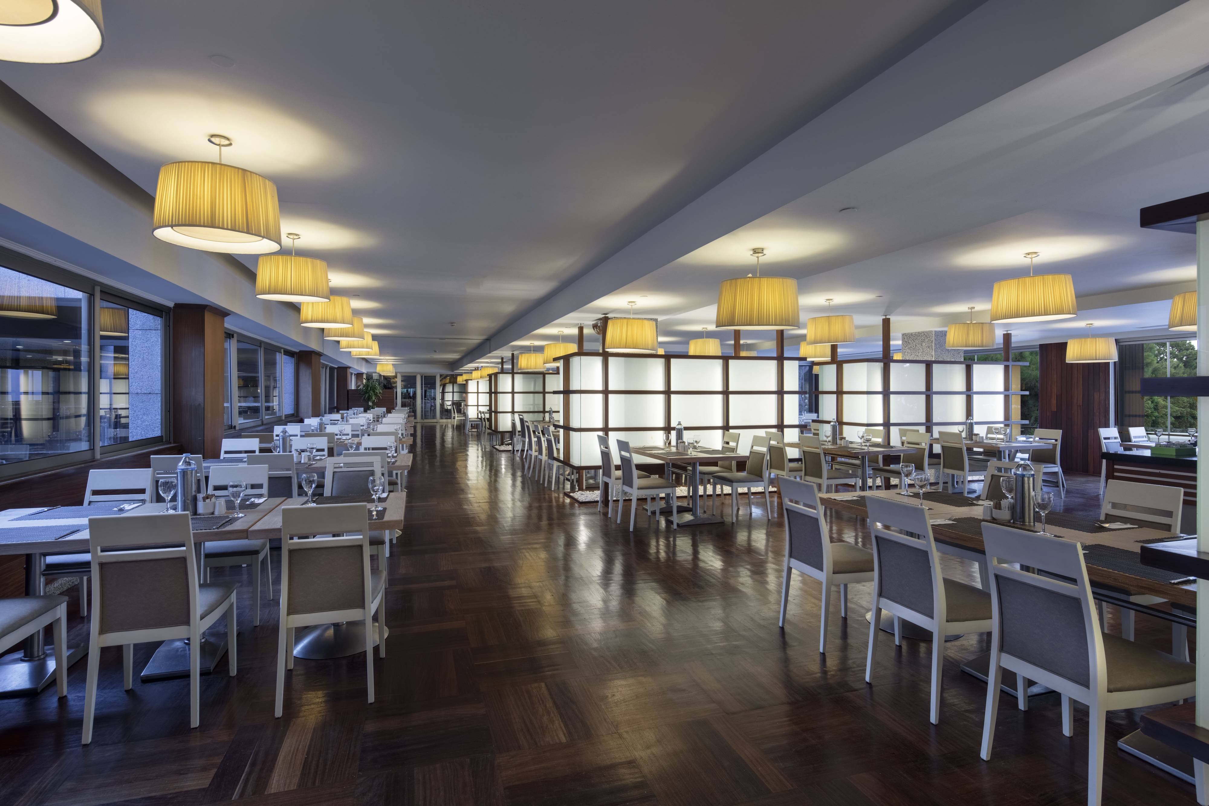 Interior view of the Veranda Main Restaurant at Rixos Sungate, featuring a long, modern dining room. It has a dark wood floor, neutral-toned seating and tables set with light linens, and is lit by a row of large, round, beige-shaded pendant lights.