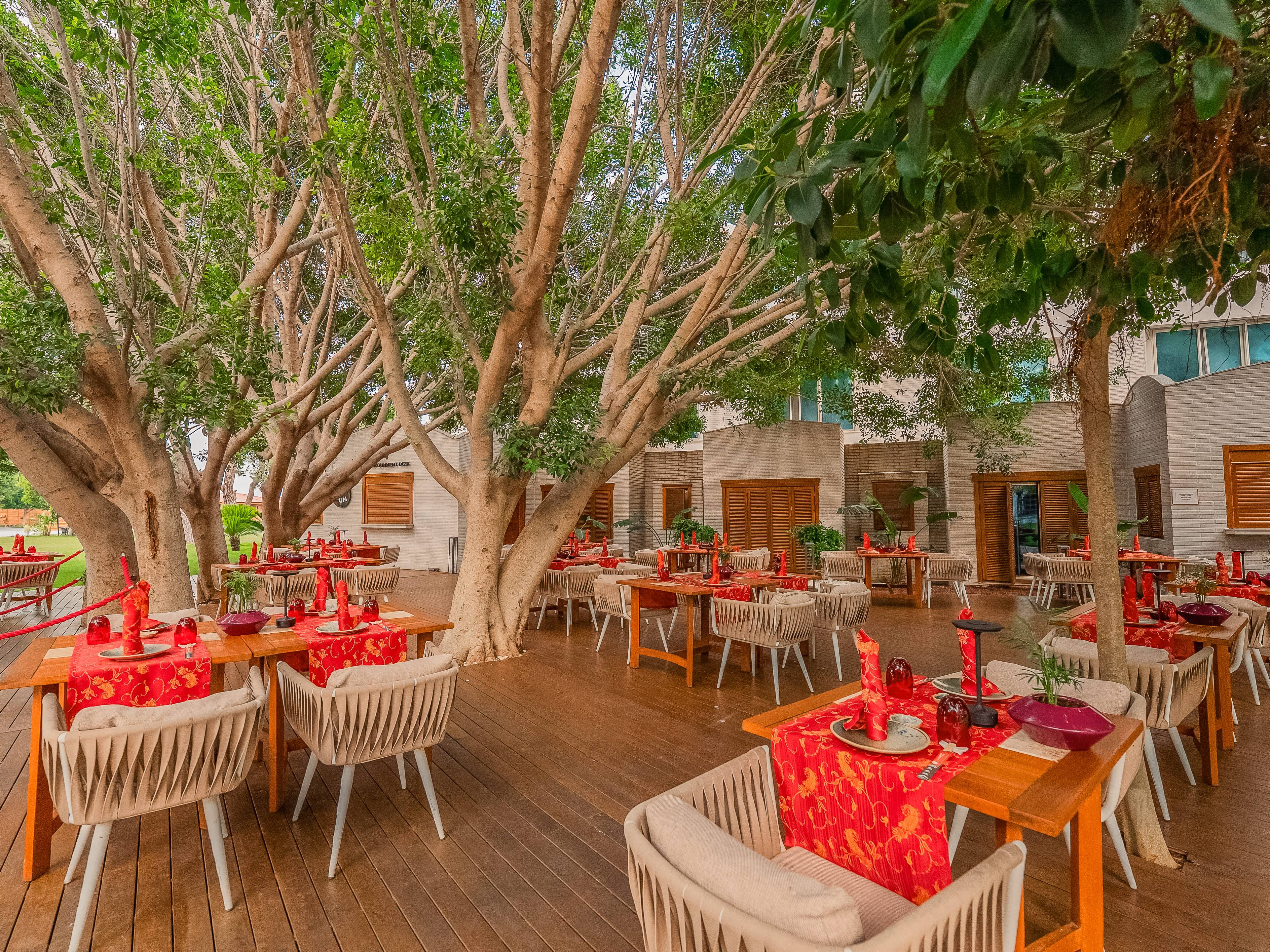 An outdoor restaurant terrace at Rixos Sungate featuring a Kuro-themed setting. The space has a dark wood deck and is shaded by large, leafy trees. Tables are set with bright red and gold patterned linens and modern, woven chairs.