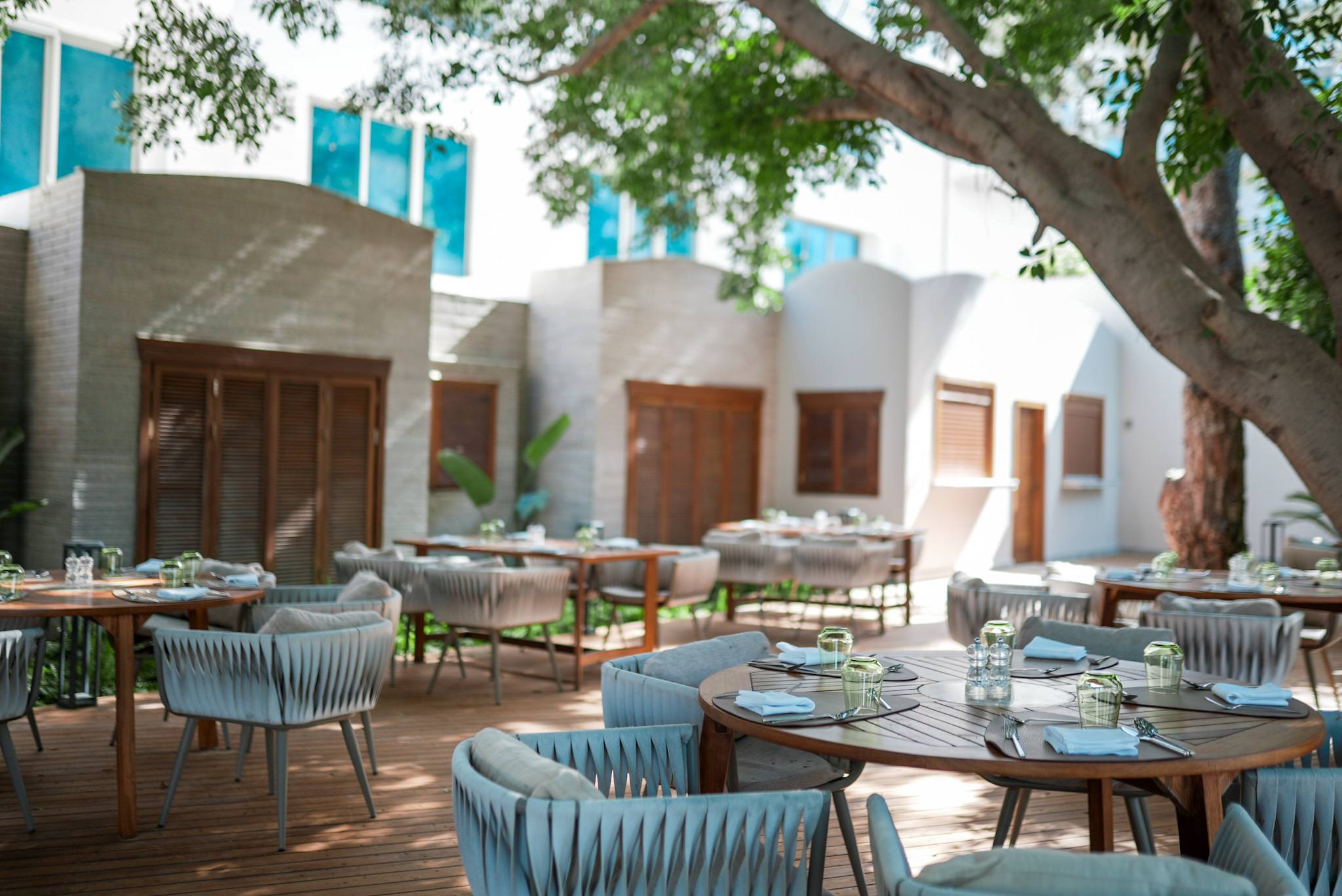 Outdoor terrace of the Noodle Service A'La Minute Restaurant at Rixos Sungate, featuring round wooden tables with modern, woven gray chairs. The space has a light wood deck, is shaded by a large tree, and is set against a neutral-toned building with wooden accents.