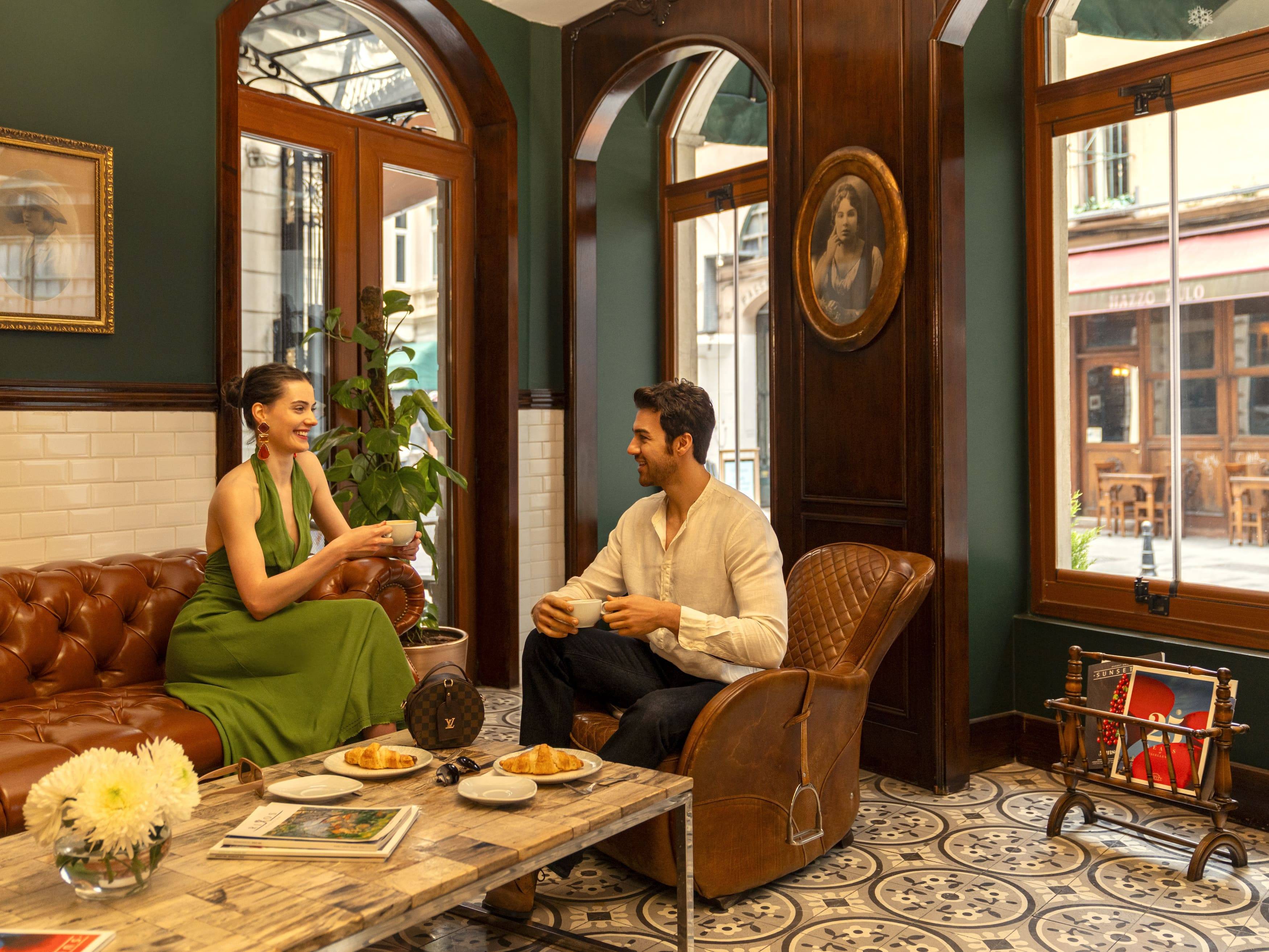 Muškarac i žena opuštaju se na smeđoj kožnoj sofi i fotelji u ugodnom CHAPELLE BISTRO & LOUNGE BAR-u s tamnozelenim zidovima u Rixos Pera Istanbulu, uživajući u kavi i pecivu.