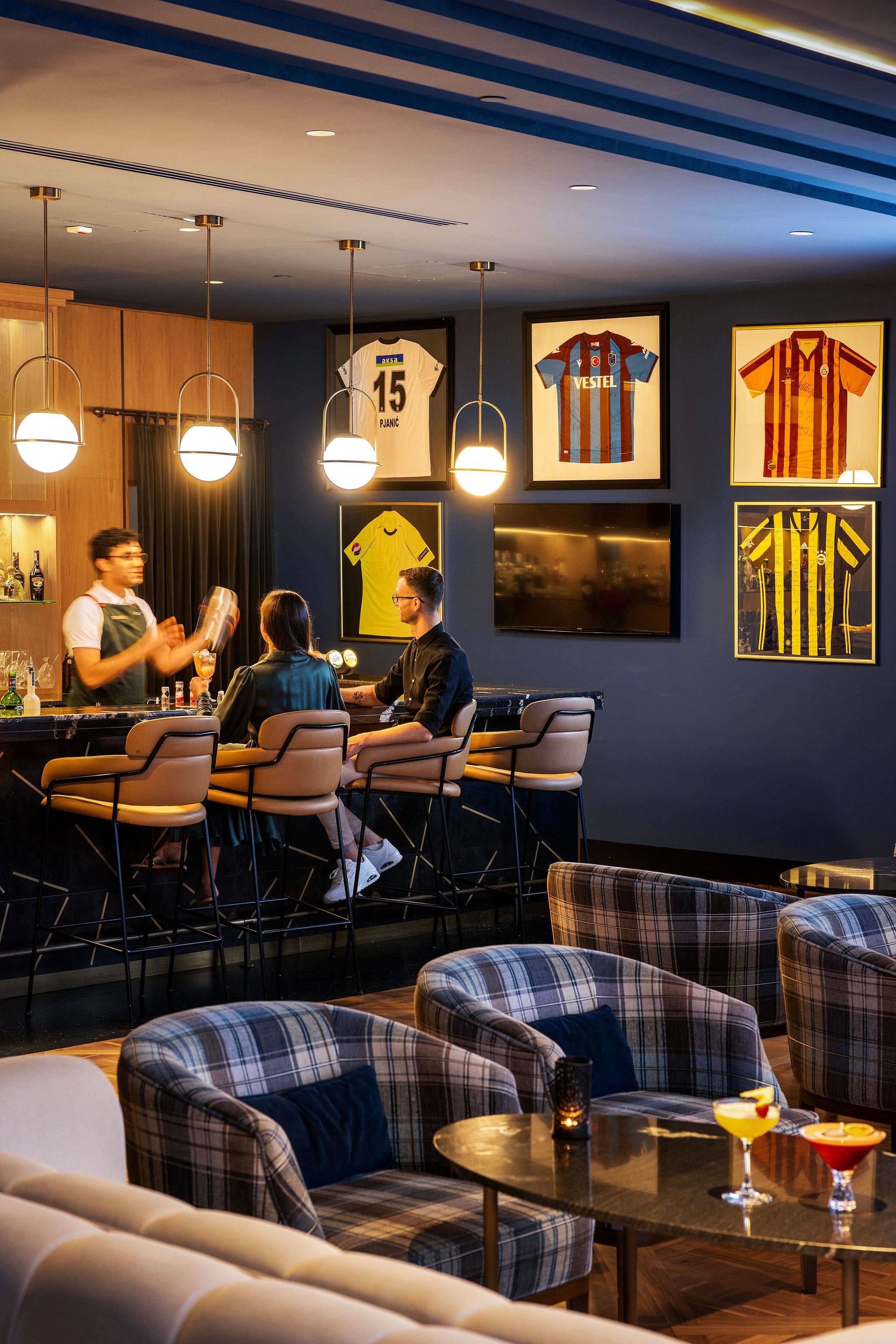 A lively vertical shot of the Sports Bar at Rixos Premium Dubrovnik. A bartender is shaking a cocktail for two guests seated at the dark marble bar, which has modern leather-backed stools. The dark blue wall is decorated with six framed sports jerseys, including one labeled "Fener" with the number 15. In the foreground, there are plaid armchairs and beige sofas with small tables holding brightly colored cocktails.