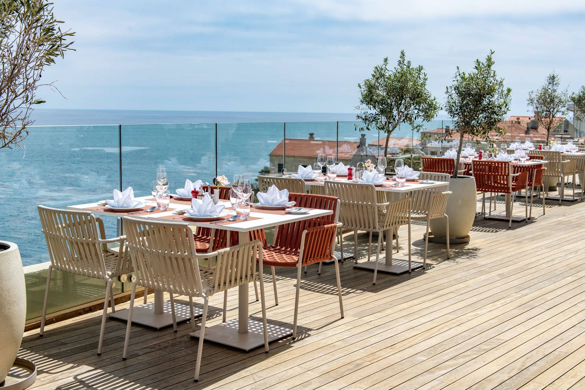 An outdoor dining terrace at the Rixos Dubrovnik overlooking the Adriatic Sea. Several tables are set with white linens, dishes, and wine glasses. The chairs are modern, in light beige and terracotta colors. The terrace has a wooden deck and a clear glass railing, offering an uninterrupted view of the deep blue sea and a cluster of terracotta-roofed buildings below.