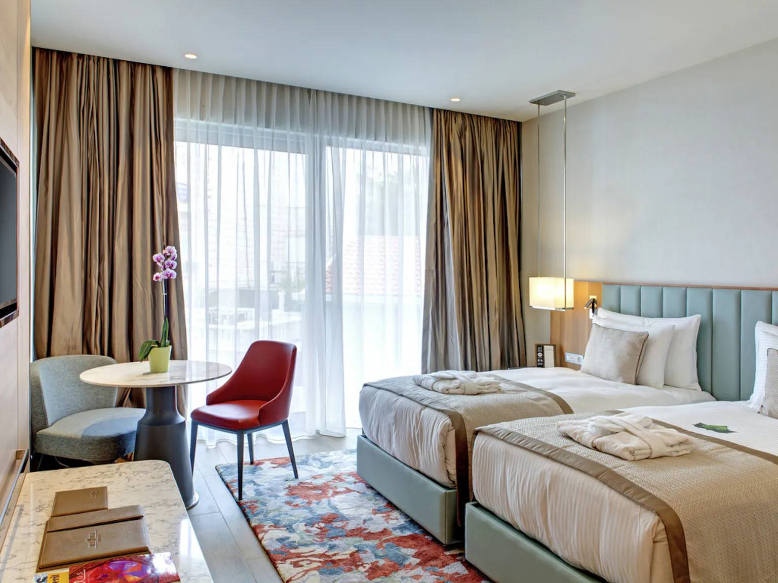 Pogled na modernu sobu s dva odvojena kreveta, malim okruglim stolom sa šarenim stolicama i velikim prozorom koji vodi na terasu