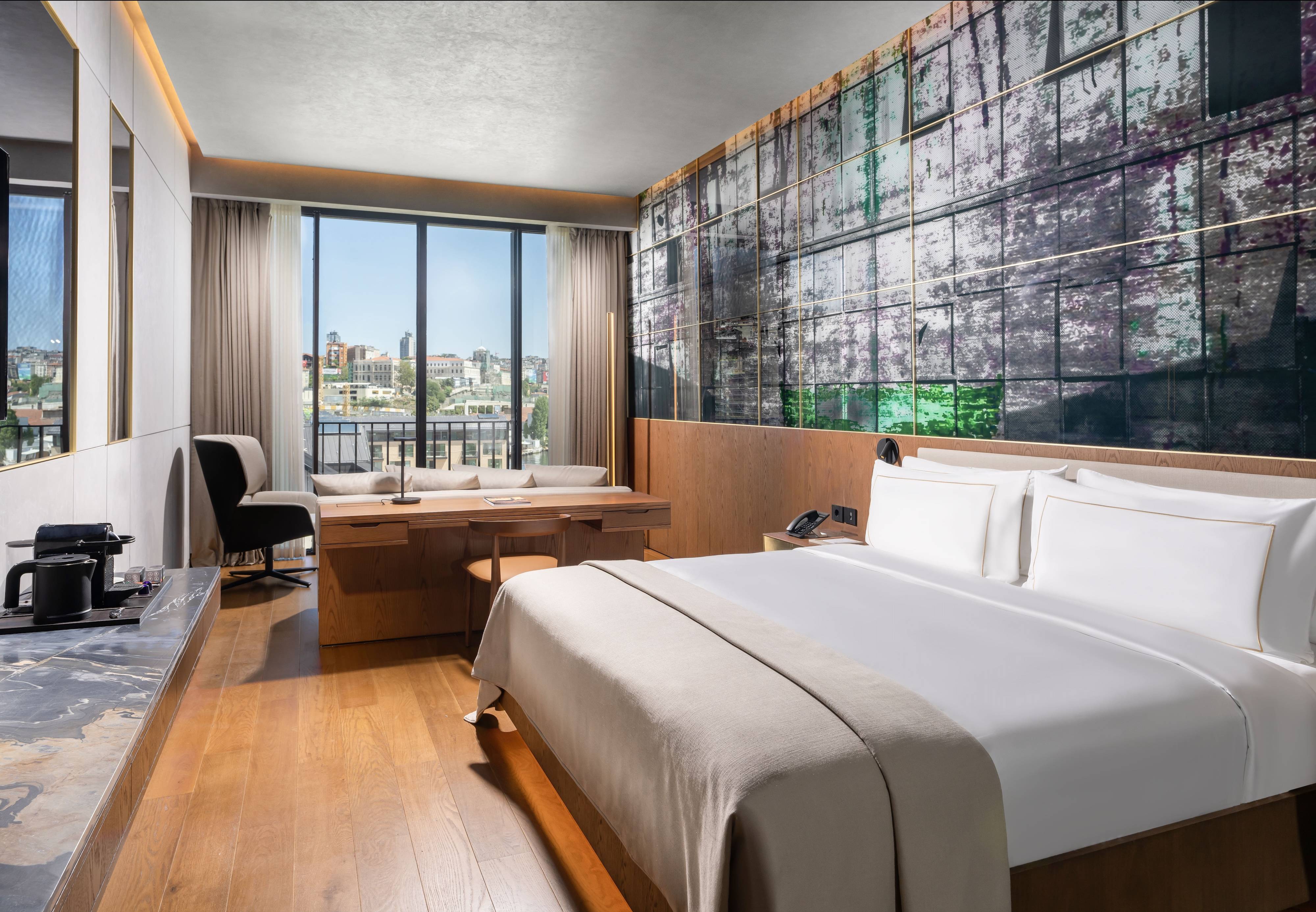 Nowoczesny pokój w hotelu Rixos Tersane Istanbul z dużym łóżkiem, biurkiem do pracy, wanną przy sięgających od podłogi do sufitu oknach z częściowym widokiem na morze, stacją kawową i dekoracyjną ścianą z abstrakcyjnymi, kolorowymi panelami.