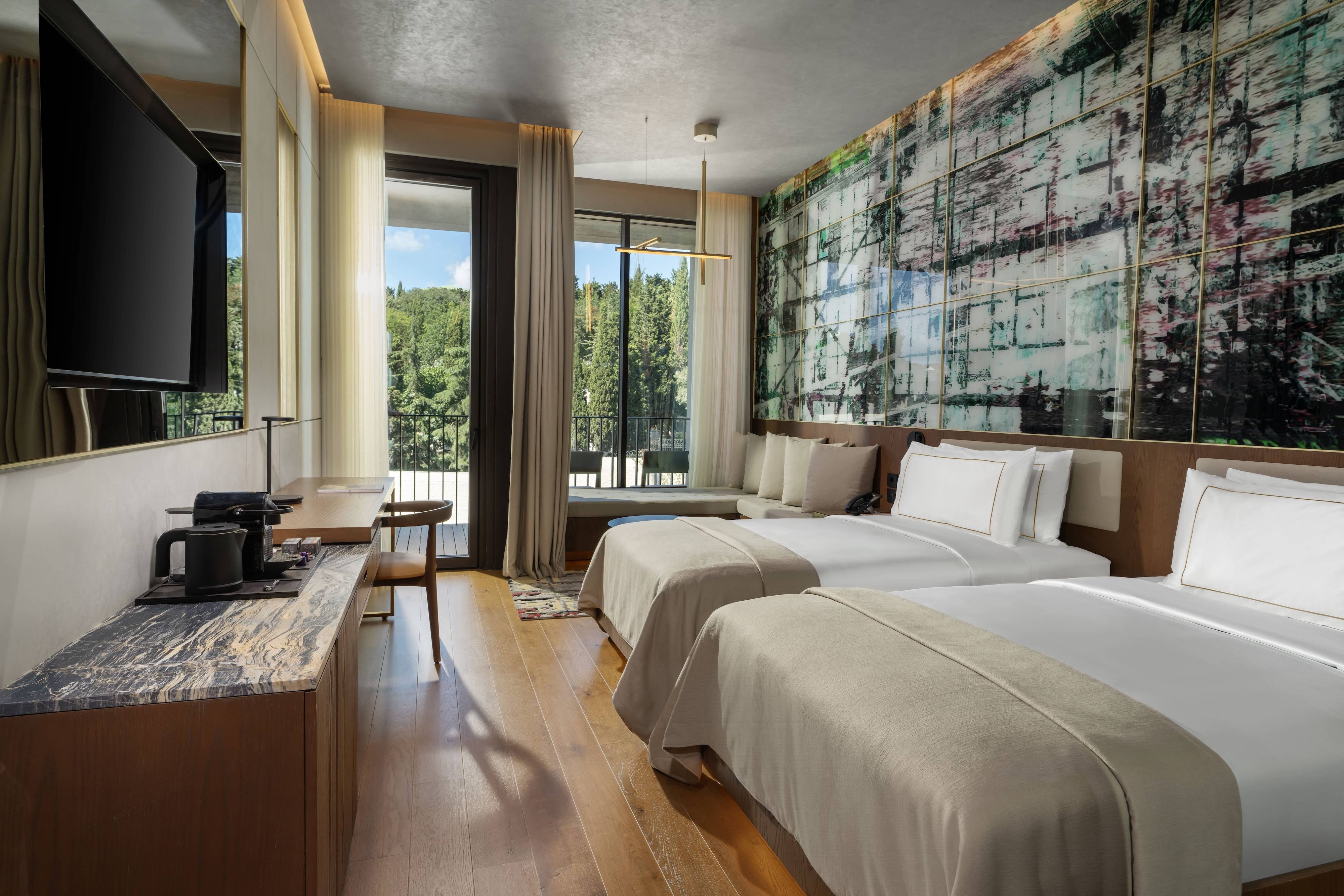 Nowoczesny pokój rodzinny typu premium z dwiema sypialniami w hotelu Rixos Tersane Istanbul z dwoma podwójnymi łóżkami, telewizorem na ścianie, biurkiem, zestawem do parzenia kawy i herbaty, dużym oknem z przezroczystymi zasłonami, balkonem z widokiem na zieleń oraz artystycznym wzorem na ścianie nad łóżkami.