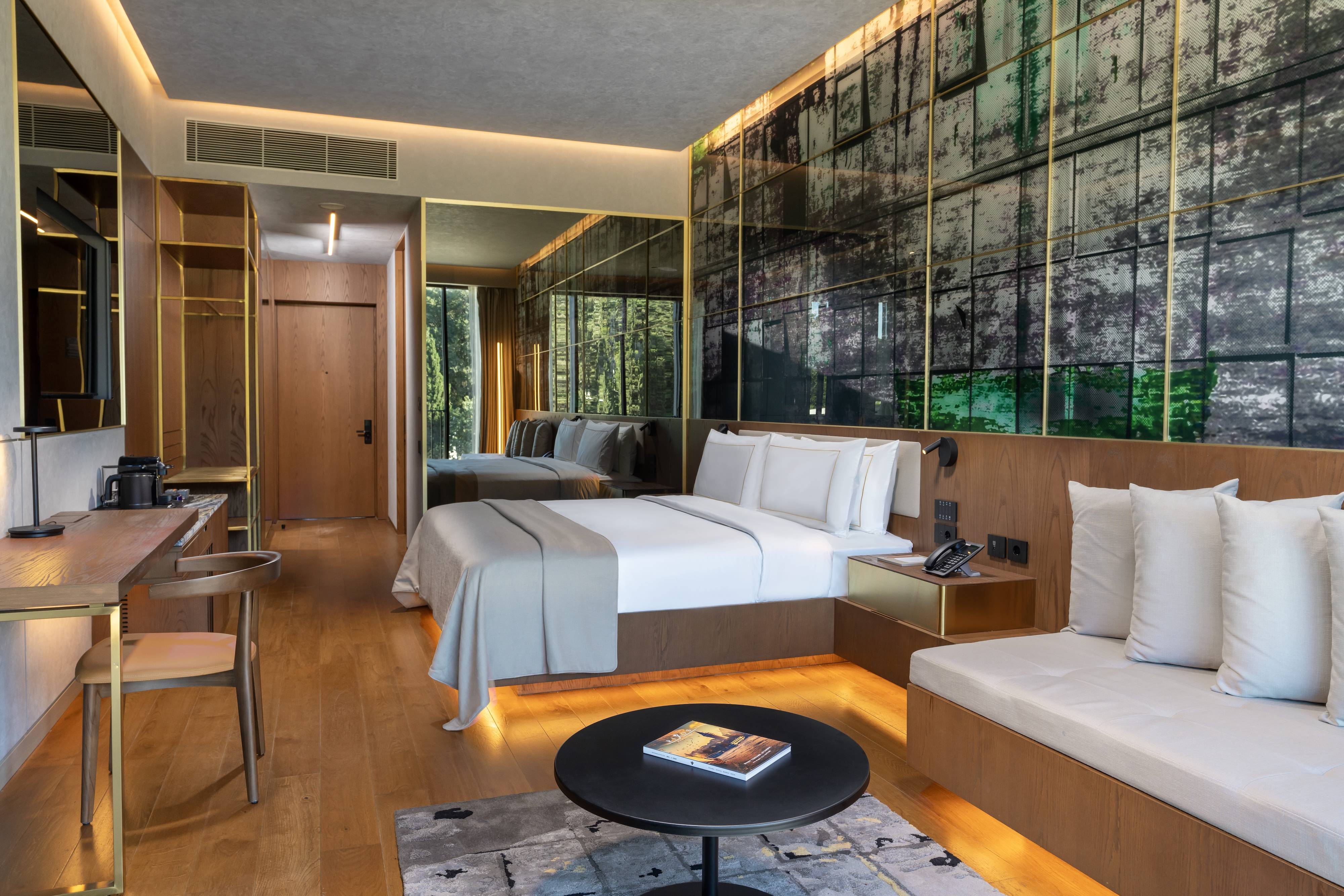 Nowoczesny pokój rodzinny Premium z dwiema sypialniami w hotelu Rixos Tersane Istanbul wyposażony jest w duże łóżko z pluszową białą pościelą, drewniane akcenty, stylową sofę, stolik kawowy z książką, duże lustra i zielone szklane panele skąpane w naturalnym świetle.