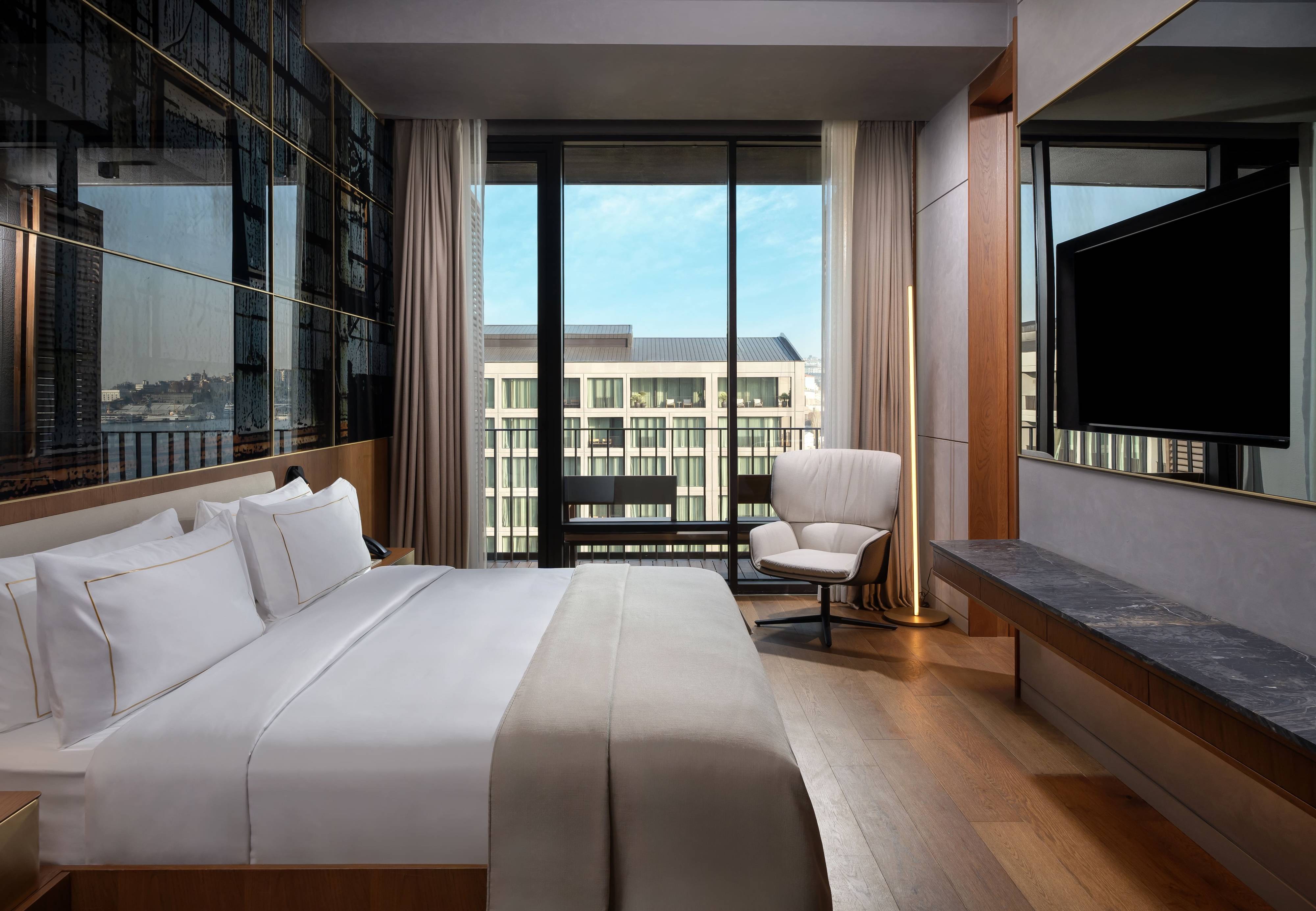 Modern hotel room in Rixos Tersane Istanbul with a large bed, white linens, cushioned chair by floor-to-ceiling windows, wall-mounted TV, wooden floors, and a view of a contemporary building outside.