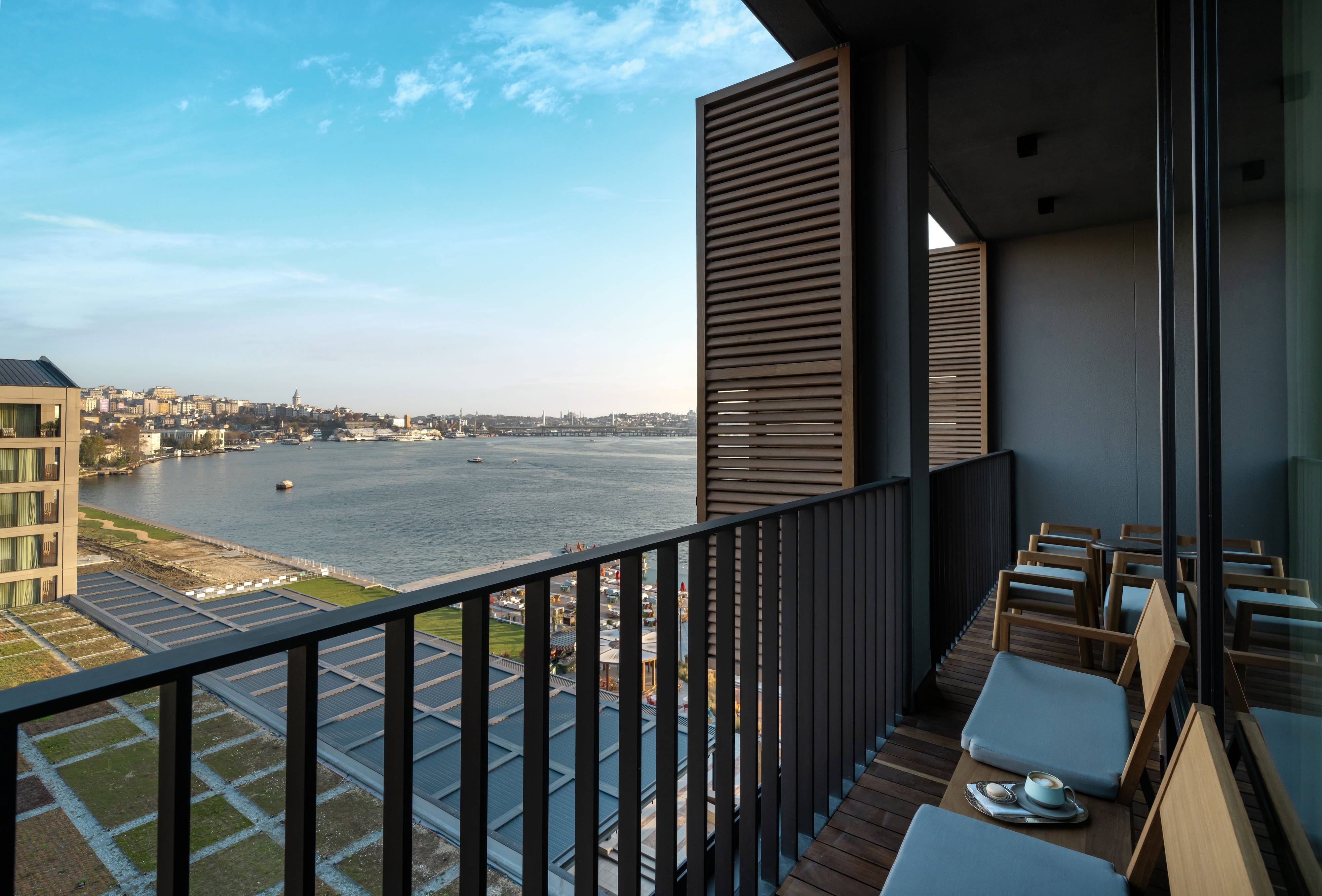 Nowoczesny balkon w dwupokojowym apartamencie typu premium suite z widokiem na morze i balkonem w hotelu Rixos Tersane Istanbul wyposażony jest w drewniane krzesła i mały stolik z widokiem na szeroką rzekę, panoramę miasta i błękitne niebo, z otwartymi okiennicami i szklanymi ścianami odzwierciedlającymi wspaniały widok.