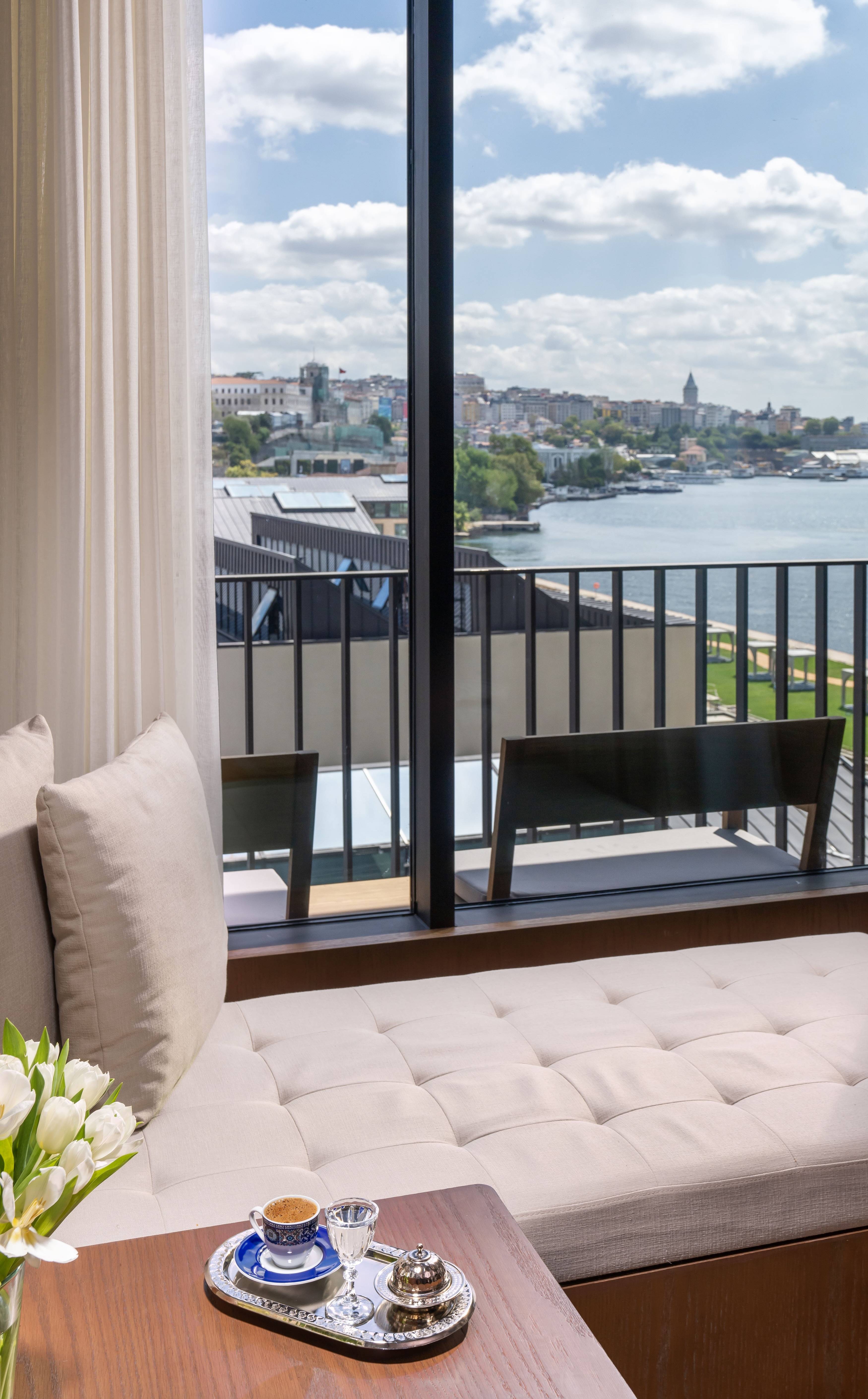 A cozy seating area by a large window in the Rixos Tersane Istanbul overlooks a river and cityscape under a partly cloudy sky. On the table, a tray with coffee, water, and white flowers in a vase completes this inviting Two Bedroom Premium Suite Partial Sea View with Balcony.