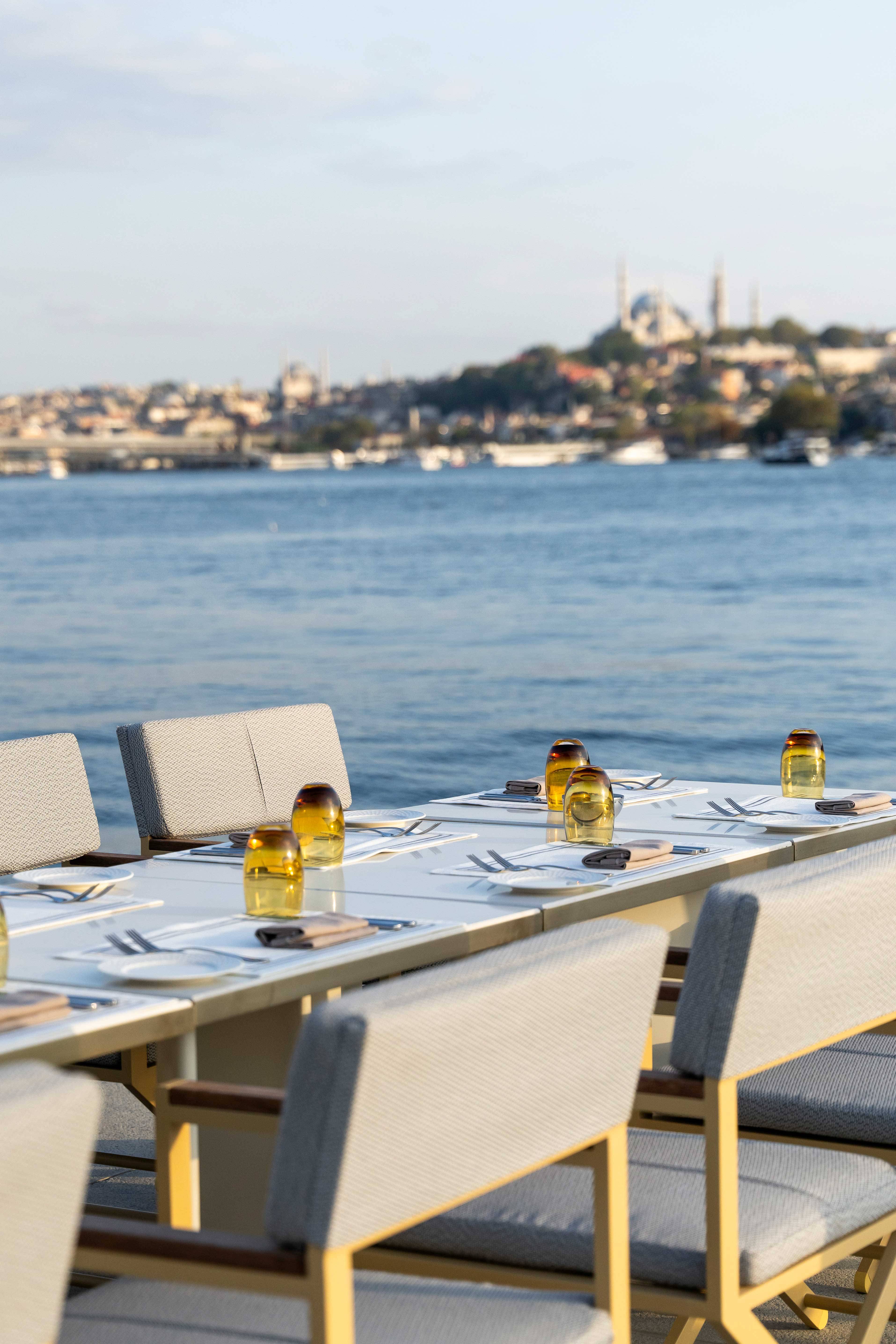 Jadalnia na świeżym powietrzu z białymi stołami i krzesłami na patio z widokiem na rzekę, zieleń i panoramę miasta z zabytkowymi budynkami i kopulastym meczetem, zaledwie kilka kroków od Velena Food Market w Rixos Tersane Istanbul pod częściowo zachmurzonym niebem.
