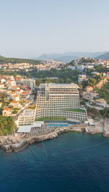 Rixos Premium Dubrovnik is a large, modern hotel with terraced balconies, set on a rocky coastline by the sea, surrounded by houses and greenery, with mountains in the background under a clear sky.