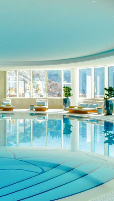 A bright, luxurious indoor swimming pool at Rixos Premium Dubrovnik features clear water, white lounge chairs with rolled towels, large potted plants, and floor-to-ceiling windows flooding the space with natural light