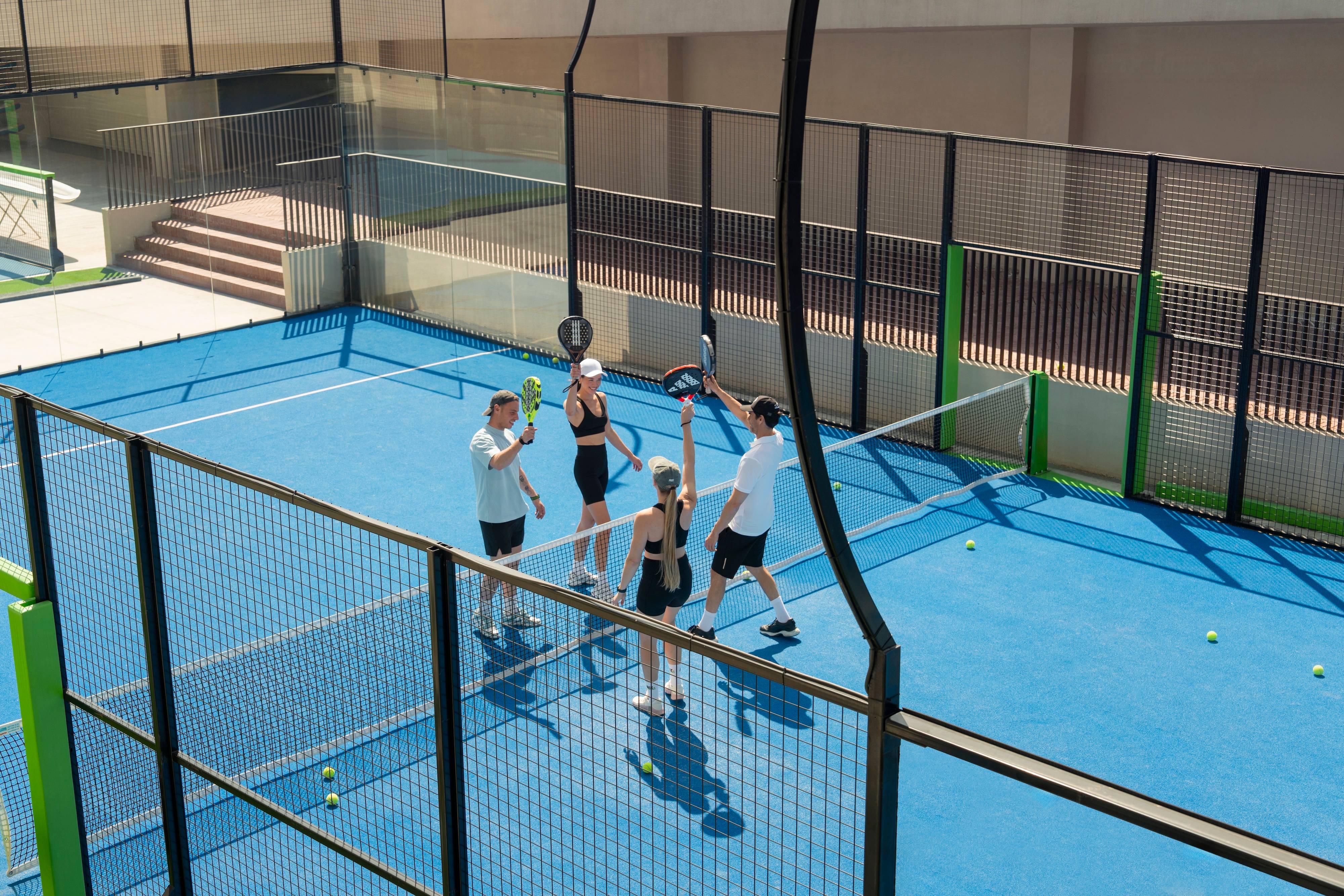 Four people on the blue padel court at Rixos Tersane cheer and high-five, holding rackets and balls. Surrounded by fences with tennis balls scattered on the ground, the mood is active and celebratory.