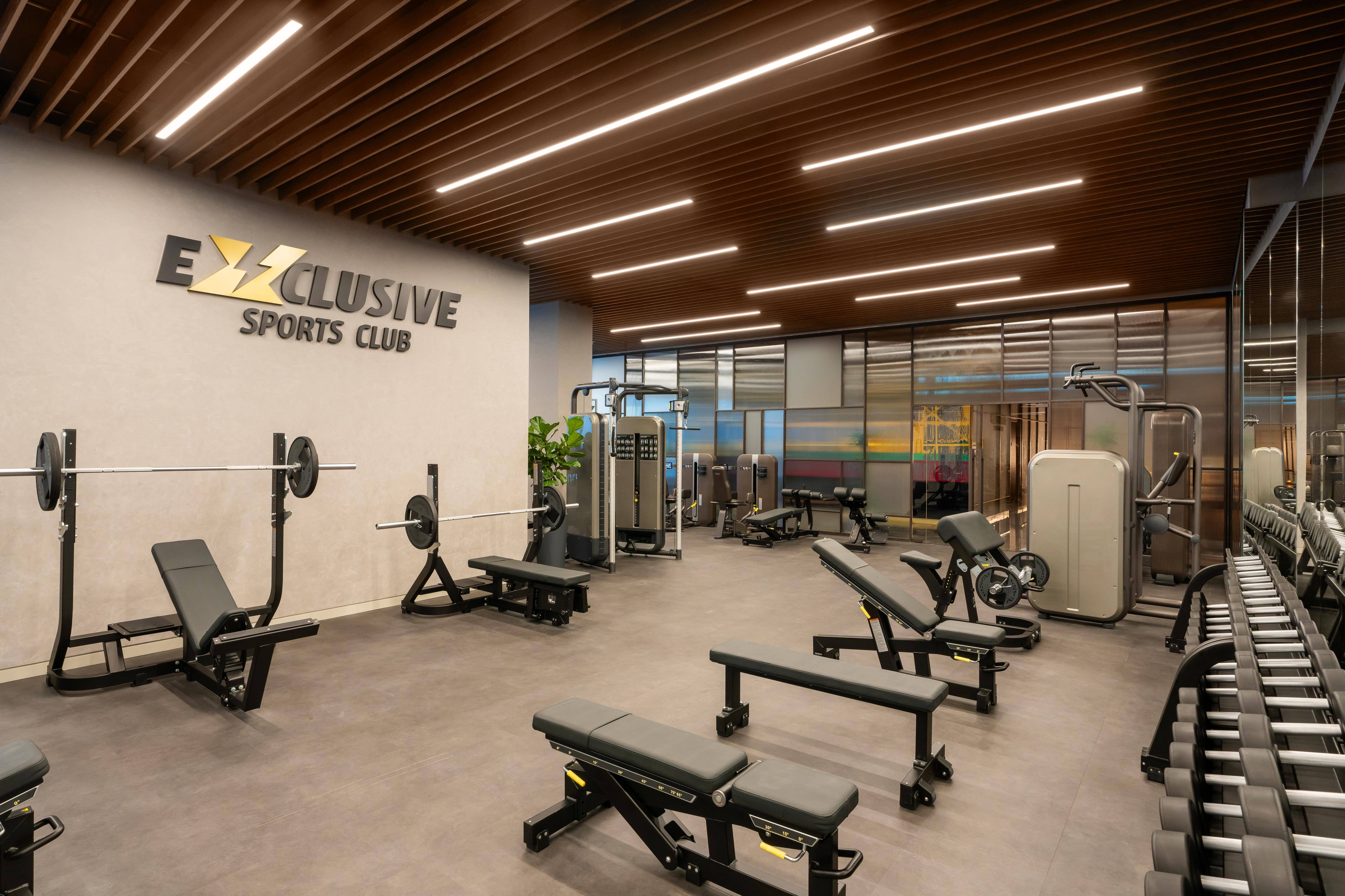 Nowoczesna siłownia w Rixos Tersane Istanbul z różnorodnym sprzętem do ćwiczeń, w tym ławkami, maszynami do ćwiczeń i hantlami. Przestrzeń ma drewniany sufit z listew, jasne oświetlenie i napis EXCLUSIVE SPORTS CLUB na ścianie.