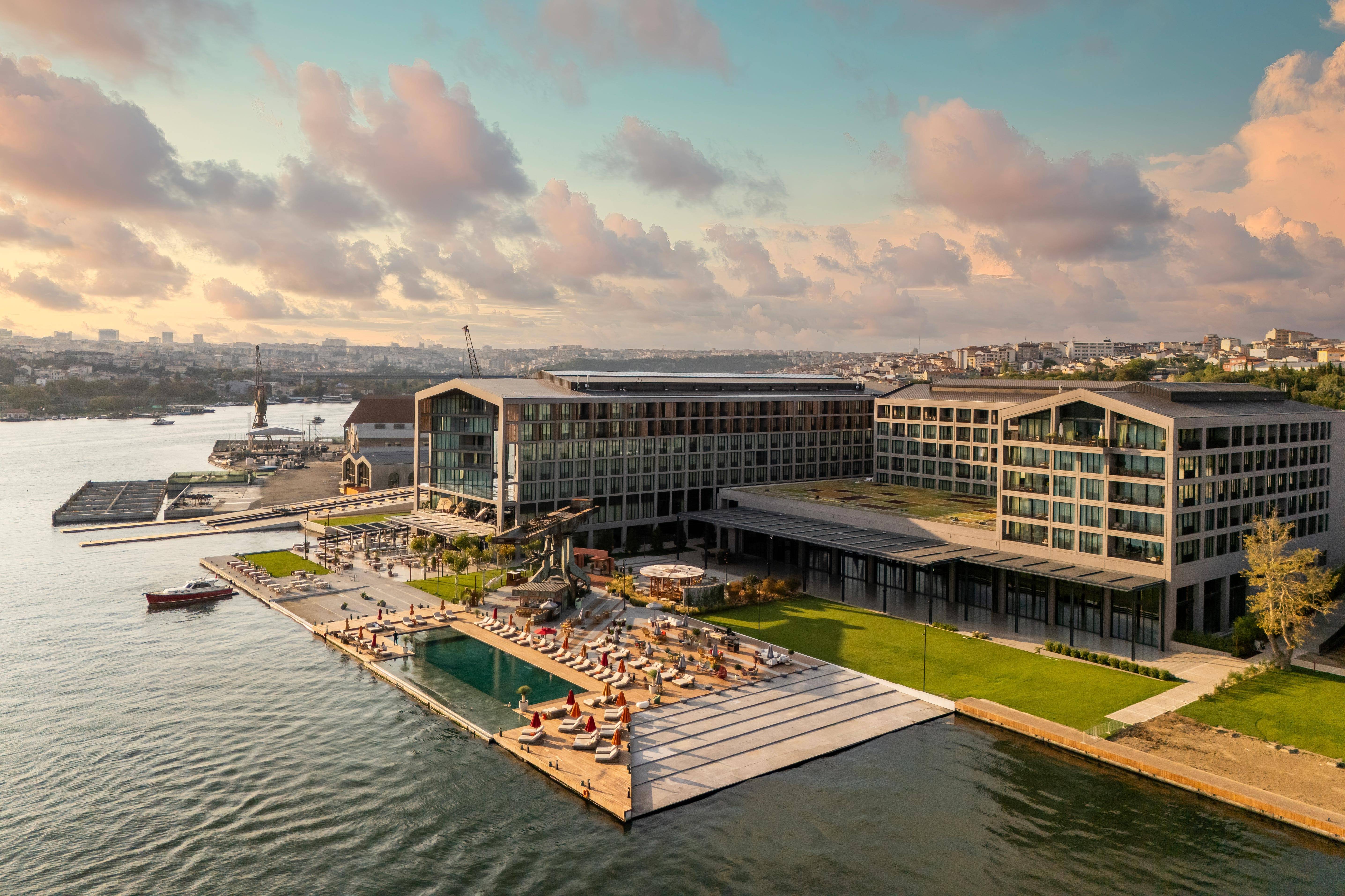 An aerial view of Rixos Tersane Istanbul overlooking the waterfront, featuring the outdoor pool, sunbathing deck, landscaped green areas, and the surrounding Golden Horn shoreline under a pastel sunset sky