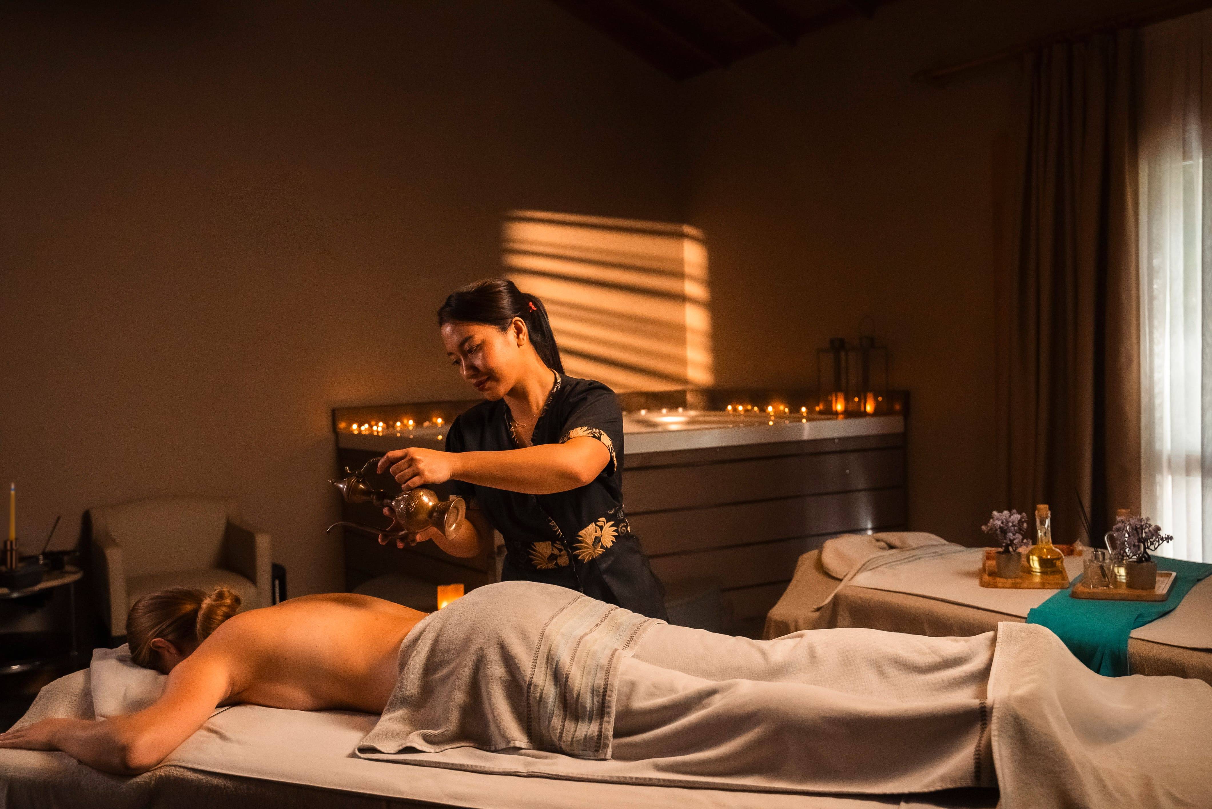 A spa therapist pouring oil onto the back of a woman lying face down on a massage table at Rixos Premium Tekirova's Anjana Spa, with ambient lighting and candles.