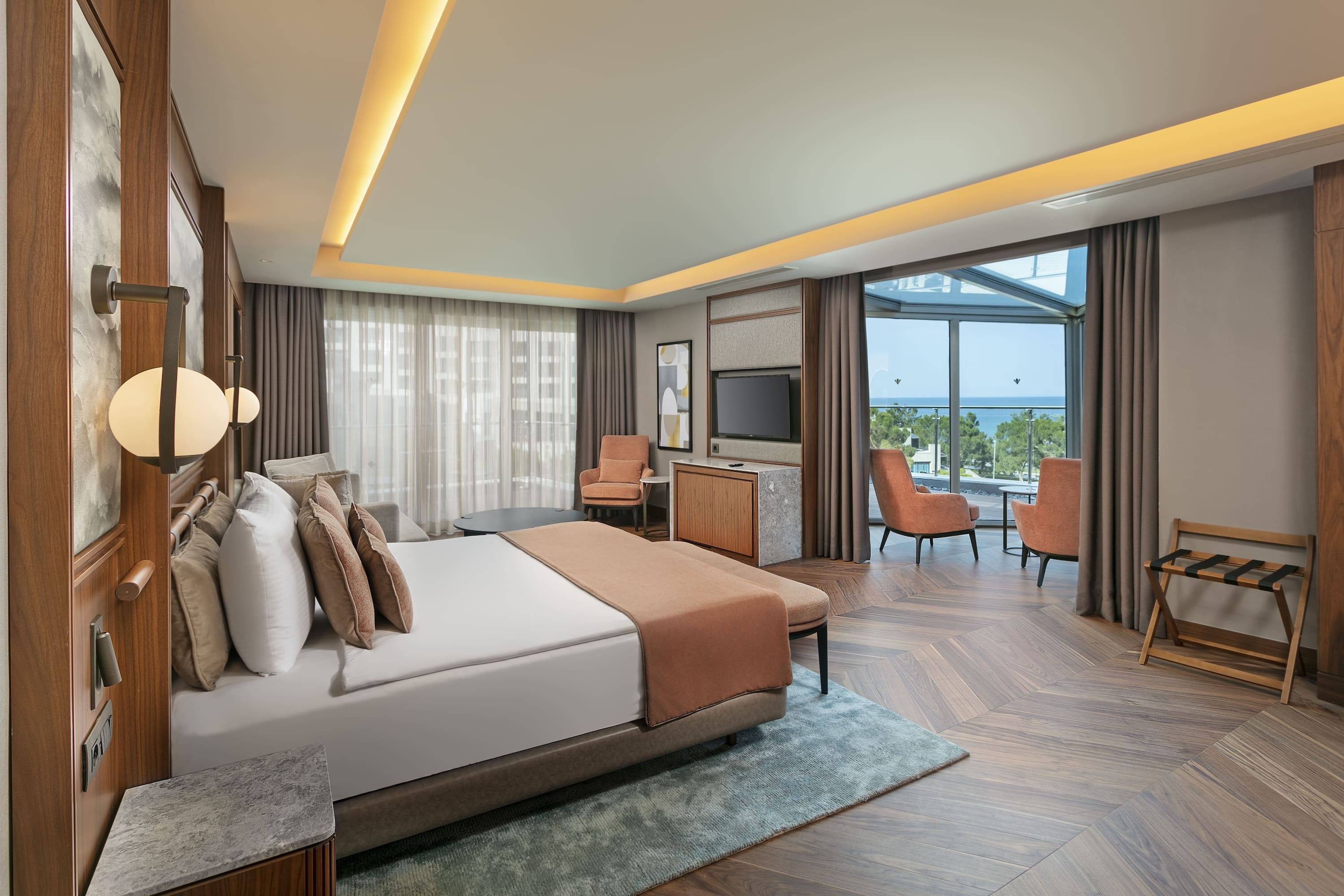 Luxurious hotel room with a large bed, herringbone wood floor, a sitting area with two orange chairs near floor-to-ceiling windows, and a sea view.