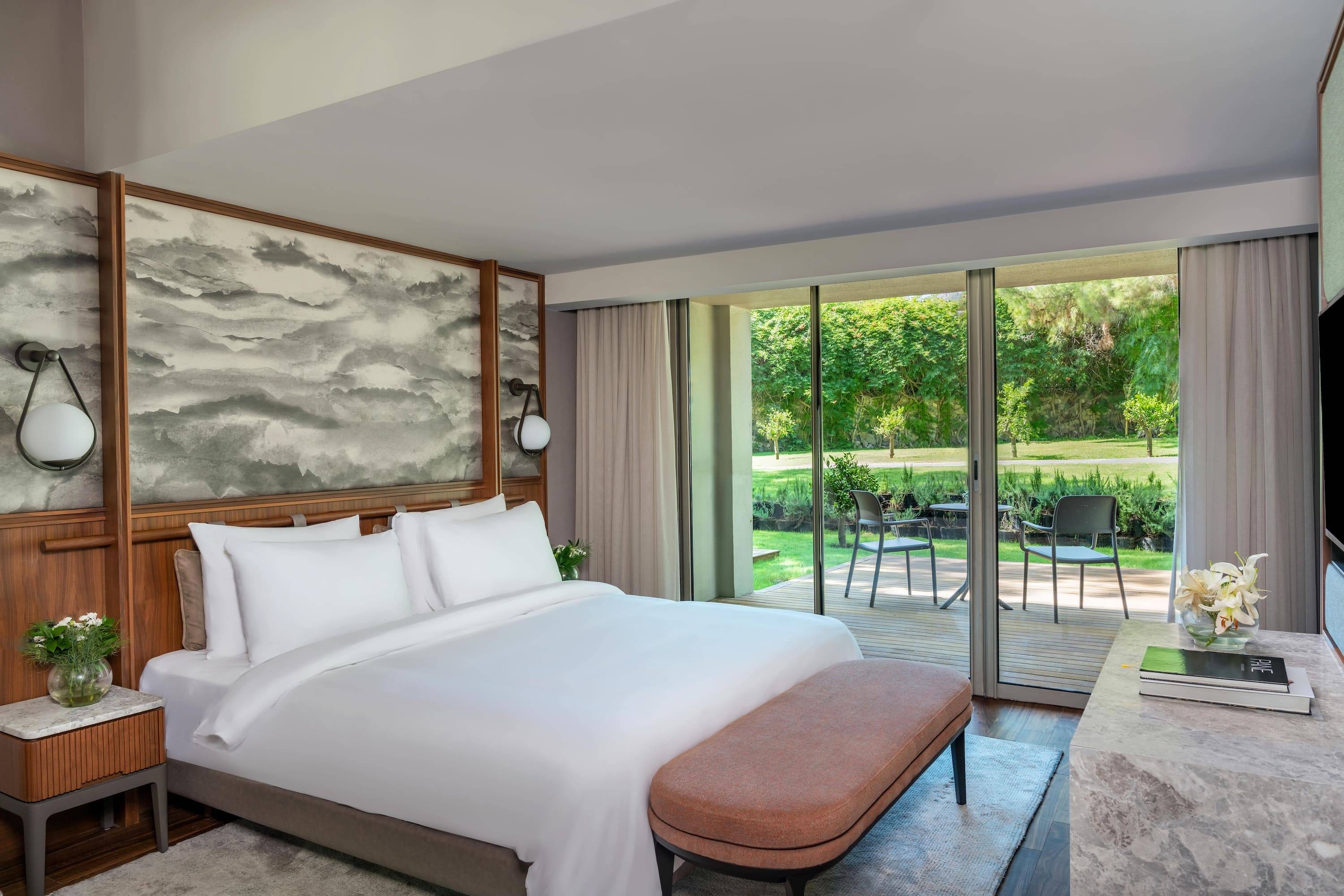 Interior view of the Marine Suite at Rixos Sungate featuring a king-size bed with a decorative grey headboard, a burnt orange bench, and floor-to-ceiling sliding glass doors opening onto a wooden terrace with a lush garden view.