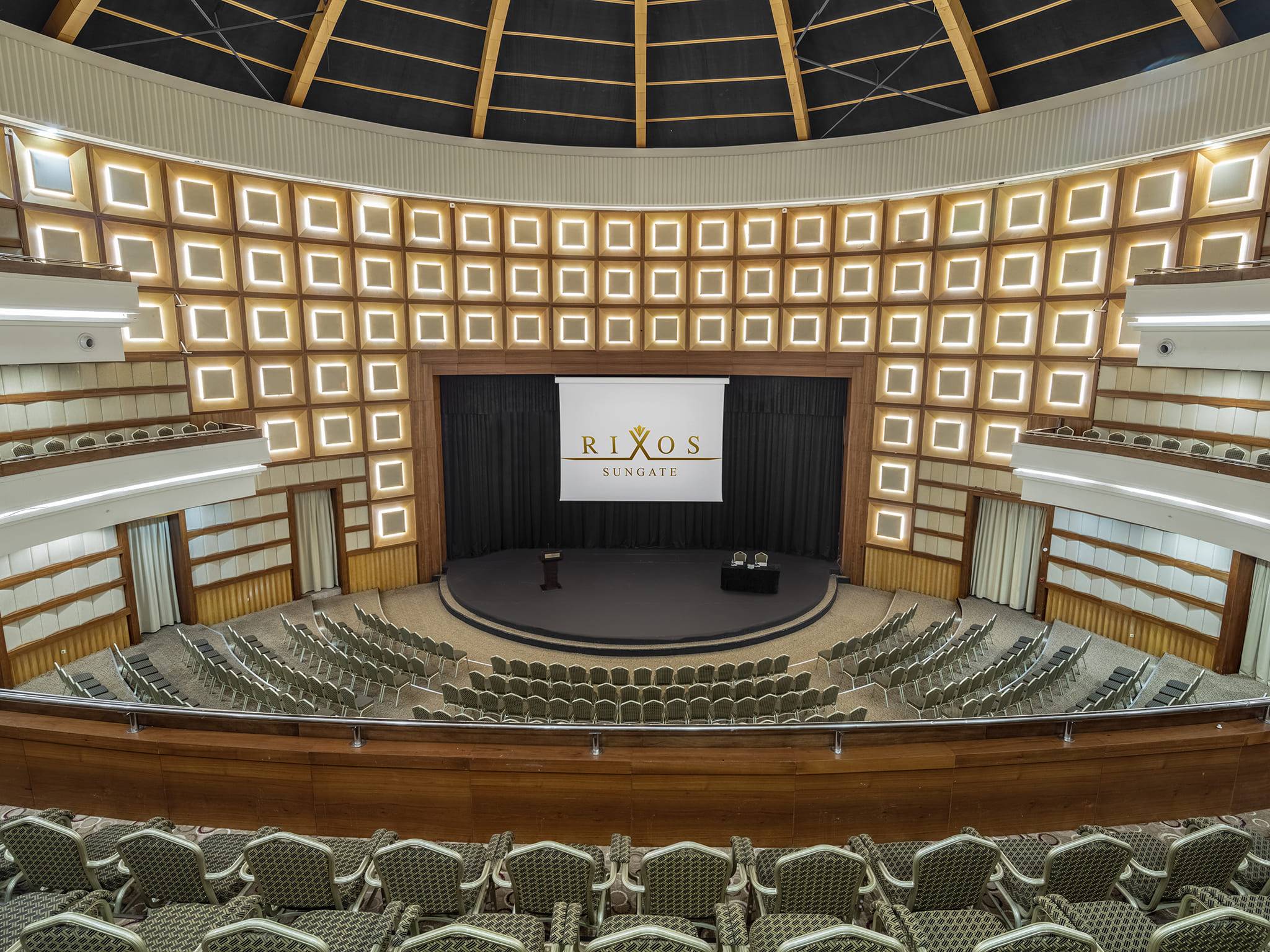 里索斯阳光门酒店的斯皮卡会议厅俯拍全景，呈现阶梯式半圆形剧场式座位布局。中央舞台清晰可见，屏幕上显示着里索斯阳光门酒店的标志。墙壁装饰着成排的背光方形灯具。此场地特别适合举办大型会议活动。