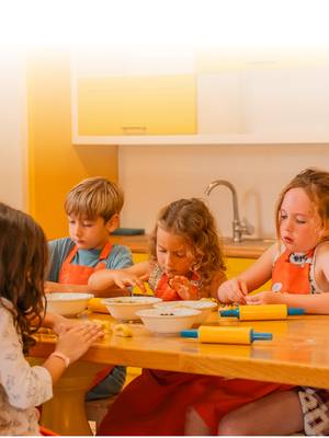 A group of four young children wearing orange aprons participating in a cooking workshop during the Winter School Break at Rixos Premium Tekirova. They are seated at a wooden table with yellow rolling pins and bowls, focused on kneading dough in a bright kitchen setting.