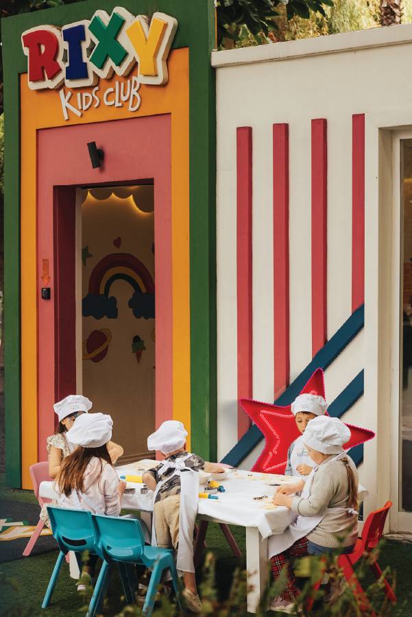A group of children wearing white chef hats and aprons participate in an outdoor cooking workshop at Rixos Downtown Antalya during the Winter School Break, seated at a table in front of the colorful Rixy Kids Club entrance