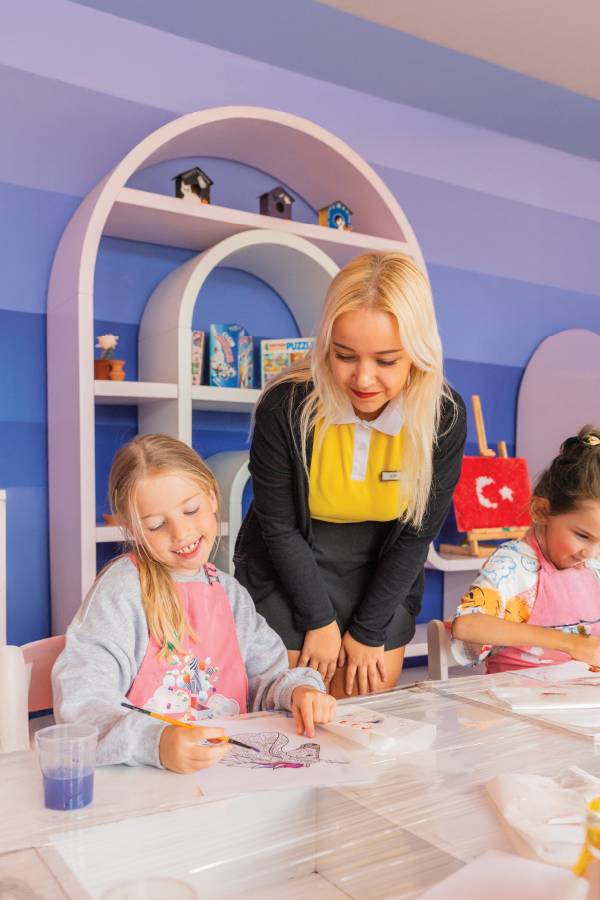 A smiling instructor supervises a young girl wearing a pink apron as she paints a unicorn drawing during an art workshop at Rixos Park Belek's Winter School Break, set in a colorful room with purple walls
