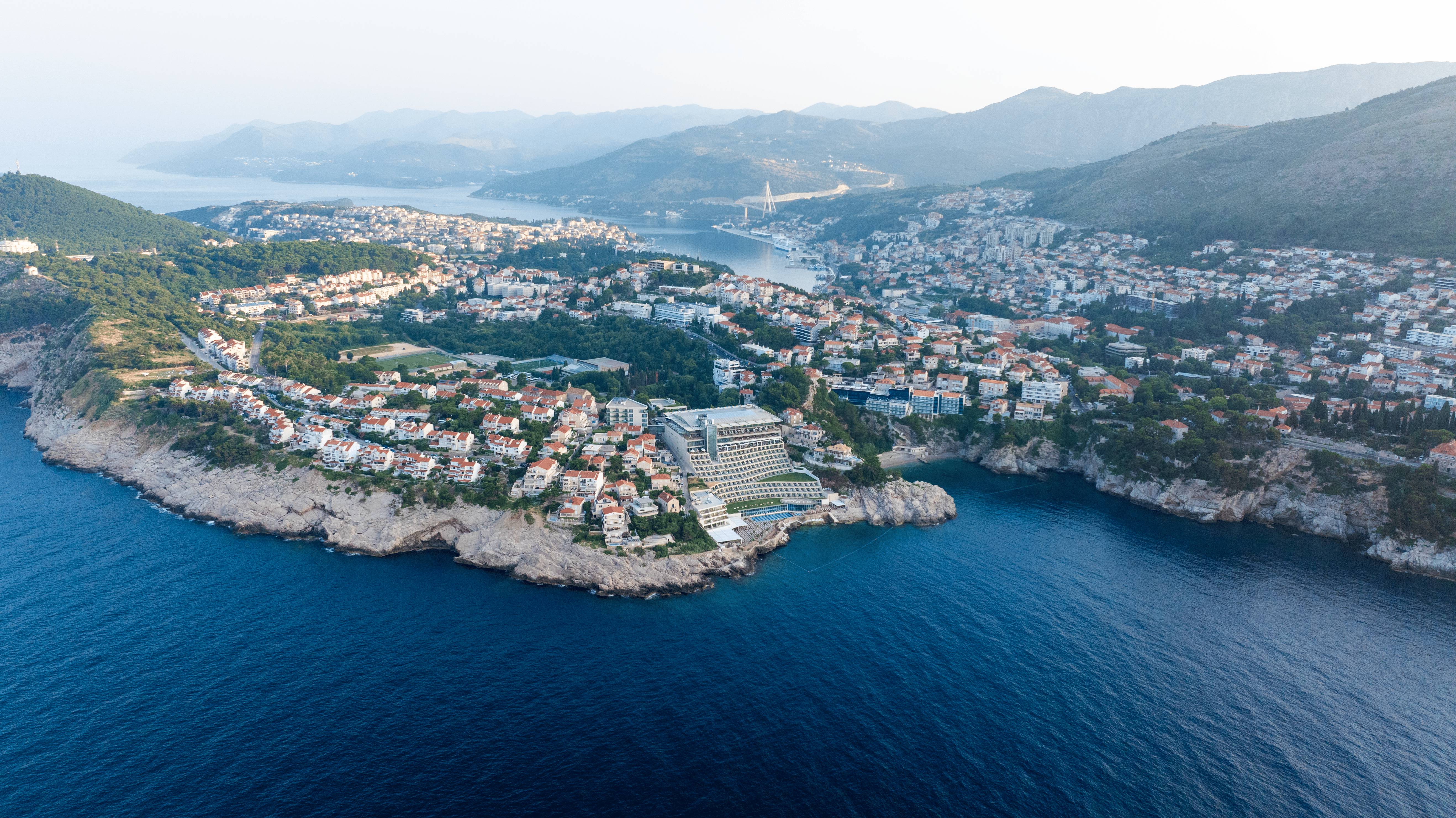 Elegantni prostor za odmor i knjižnica u Rixos Premium Dubrovniku s policama za knjige u tirkiznoj boji od poda do stropa i modernim crnim kaminom. U sredini visi velika apstraktna slika s preljevom od žute do zelene boje. U prvom planu, crne žičane fotelje s tirkiznim jastucima okružuju niski drveni stolić za kavu prepun šarenih umjetničkih knjiga." Sažeto: "Knjižnica u predvorju Rixos Premium Dubrovnik u tirkiznoj boji s kaminom i modernom umjetnošću."