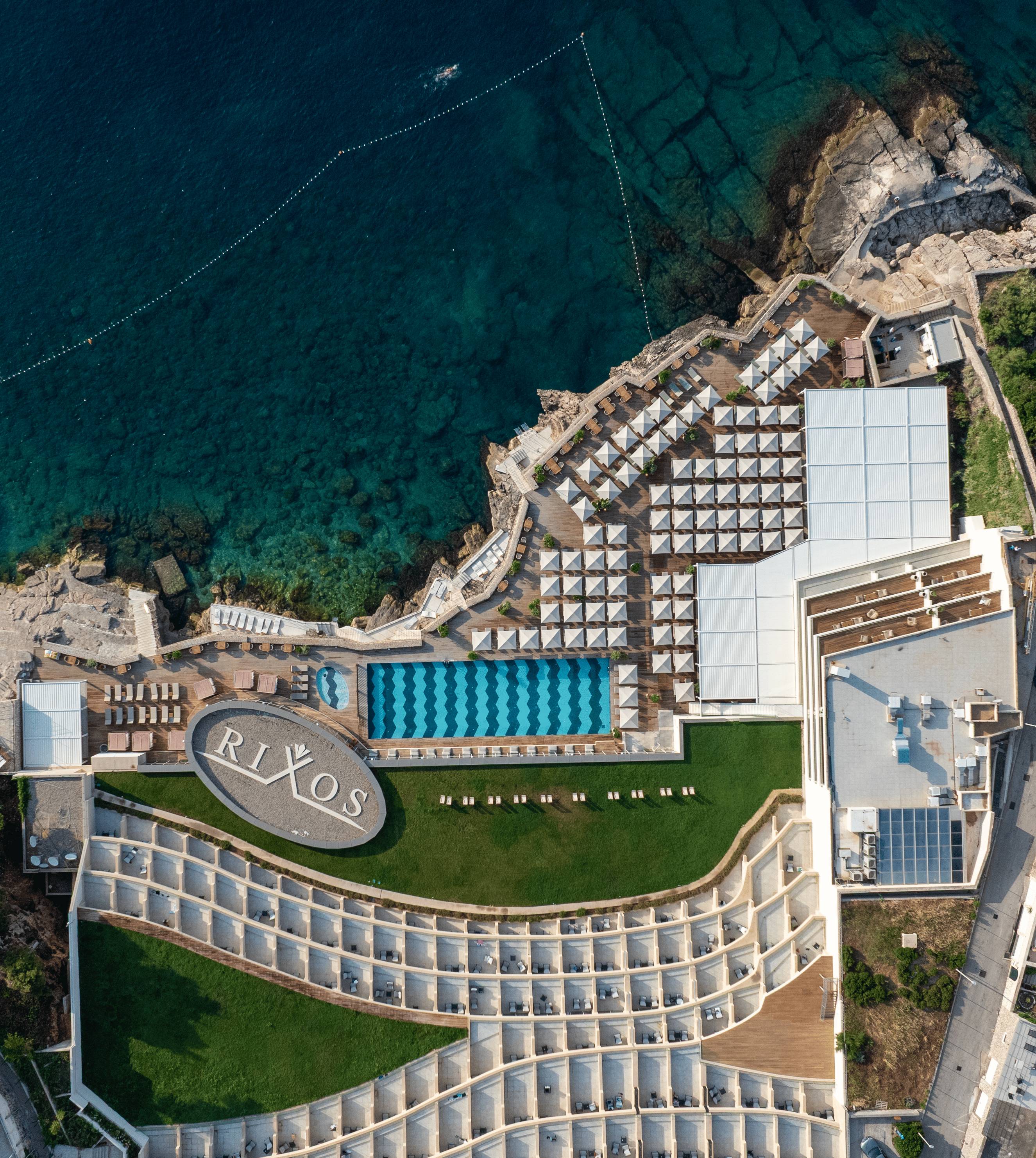Snimka iz zraka iz ptičje perspektive na odmaralište Rixos Premium Dubrovnik snimljena dronom. Slika prikazuje vanjski bazen hotela s uzorkom plavih cik-cak pločica, drvenu terasu za sunčanje ispunjenu bijelim suncobranima i veliki ovalni krajobrazni element s natpisom 'RIXOS'. Odmaralište se nalazi na rubu stjenovitog grebena s pogledom na duboko plavoplavo Jadransko more.