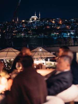 Vue nocturne de la silhouette illuminée d'Istanbul et d'une mosquée historique de l'autre côté de l'eau, depuis la terrasse extérieure du Mykorini au Rixos Tersane Istanbul, avec des clients assis au premier plan.