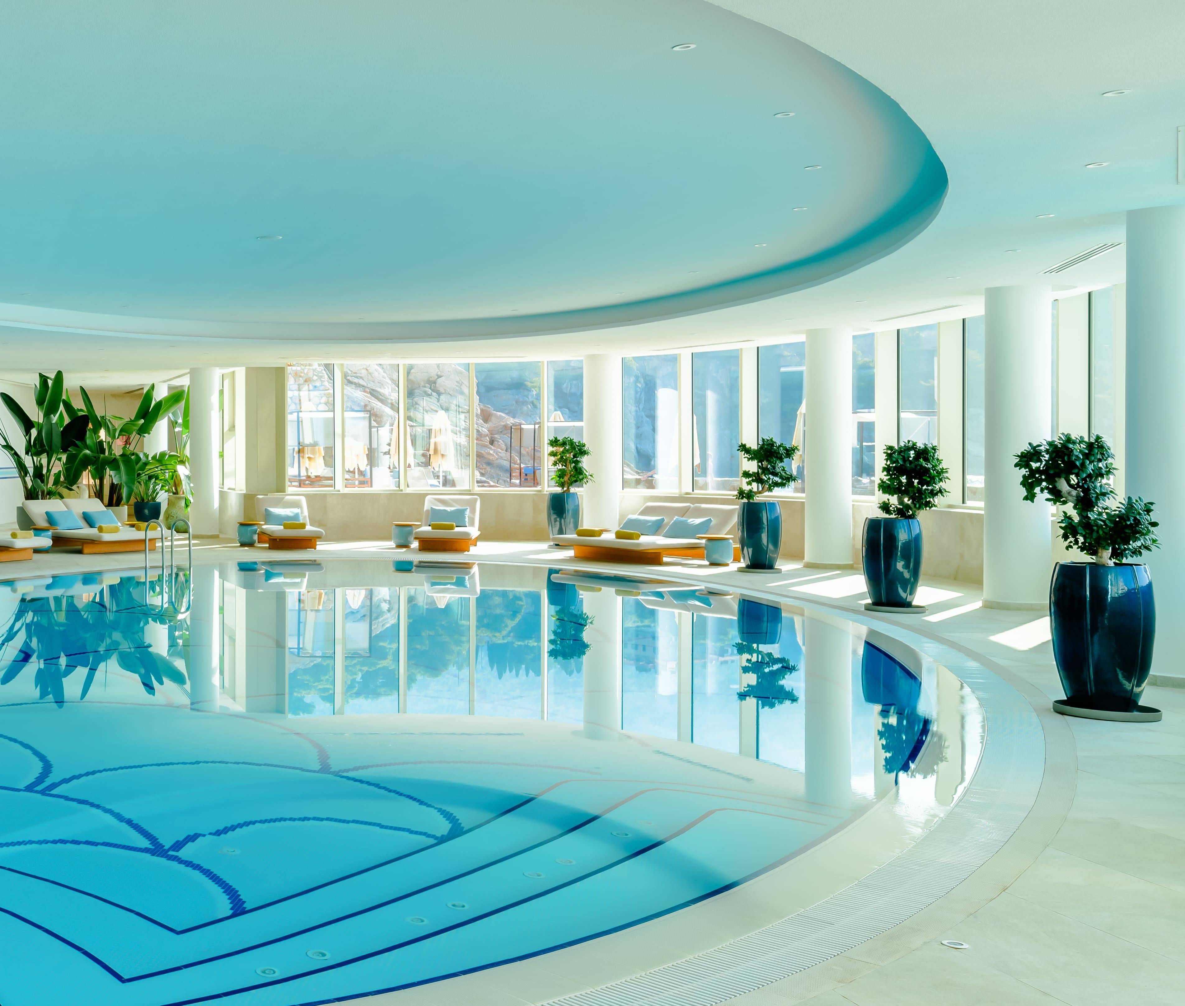 Svijetao i prostran unutarnji bazen u Rixos Premium Dubrovniku. Prostor ima prozore od poda do stropa s prirodnim svjetlom, velike bijele stupove i zakrivljeni dizajn bazena. Stabla fikusa u teglama i ležaljke sa žutim jastucima krase bazen.