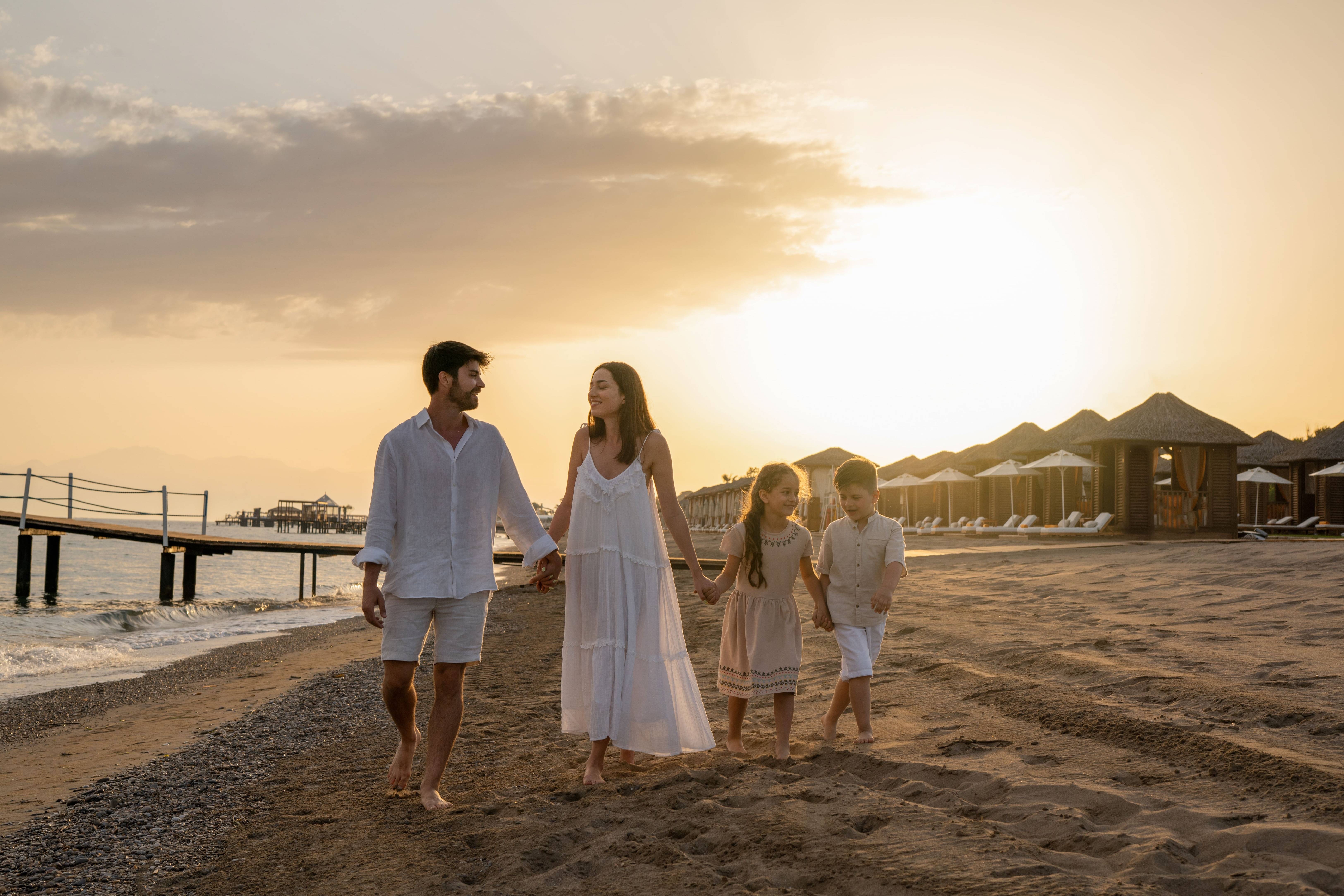 Une famille de quatre personnes marchant main dans la main au coucher du soleil sur la plage de sable du Rixos Premium Belek, avec la jetée et les cabanes privées en arrière-plan.