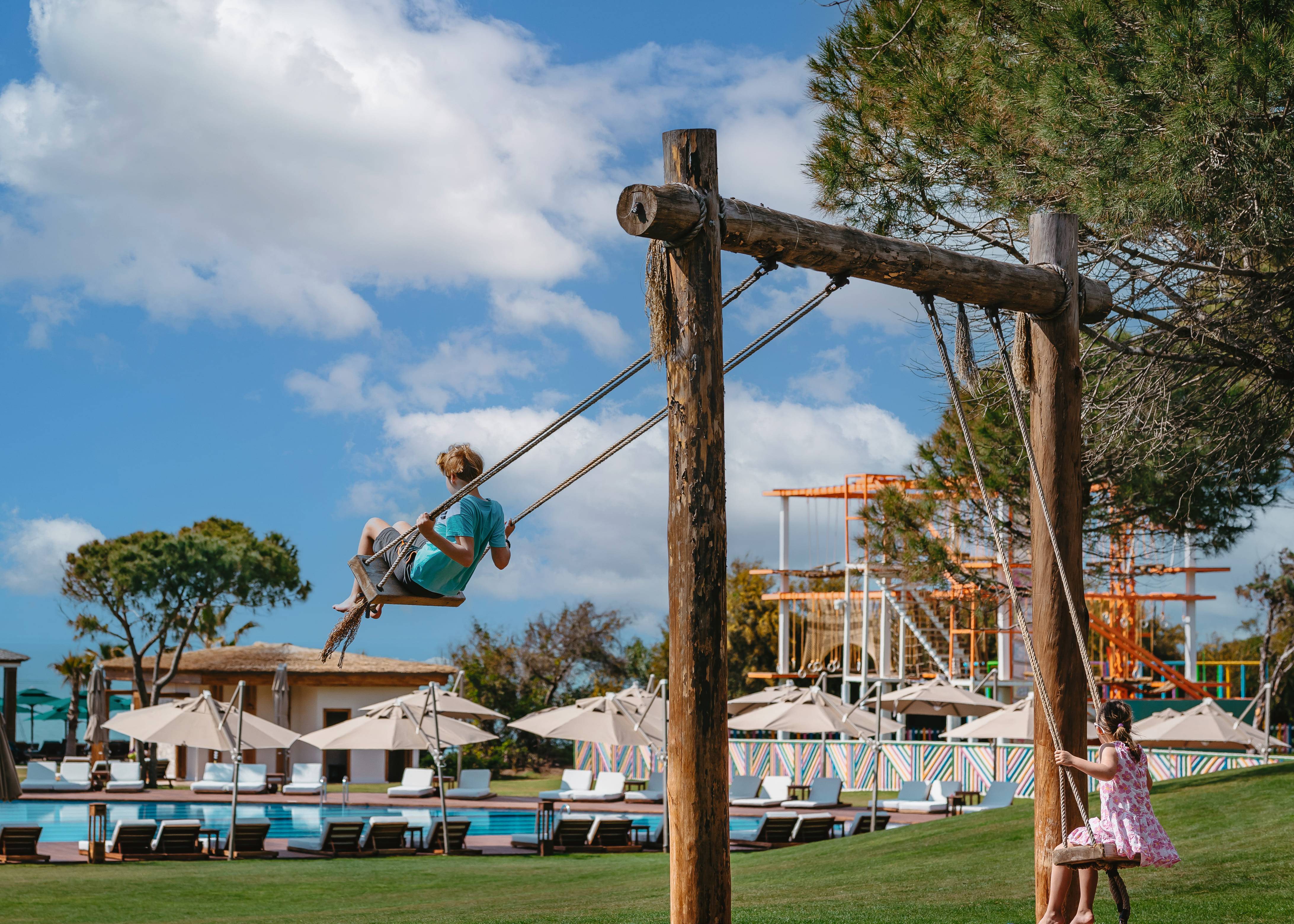 Children playing on a rustic wooden swing set on a grassy lawn, with the main swimming pool and umbrellas in the background at Rixos Premium Belek