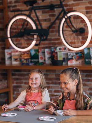 فتاتان صغيرتان مبتسمتان ترتديان مآزر وردية اللون ترسمان أوعية خزفية خلال ورشة عمل فنية في نادي Rixos Premium Belek Rixy Kids Club. تظهر في الخلفية جدار من الطوب الريفي مزين بدراجة ذات عجلات مضيئة ورف مليء بألعاب الطاولة.