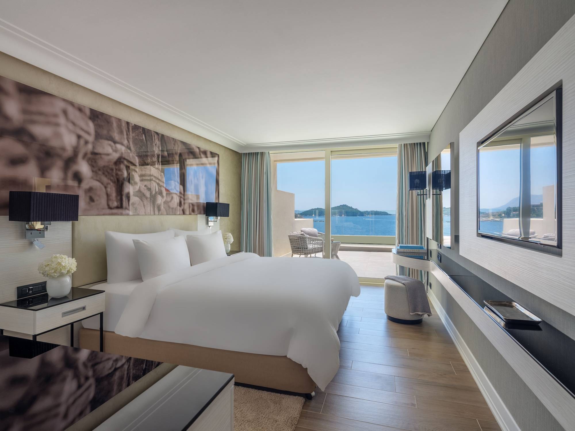 Svijetla i moderna Superior soba s pogledom na more u hotelu Rixos Premium Dubrovnik sadrži king size krevet s bijelom posteljinom, veliki arhitektonski zidni mural i svijetle drvene podove. Klizna staklena vrata od poda do stropa otvaraju se na balkon sa sjedećom garniturom, s kojeg se pruža panoramski pogled na plavo Jadransko more i udaljeni otok.