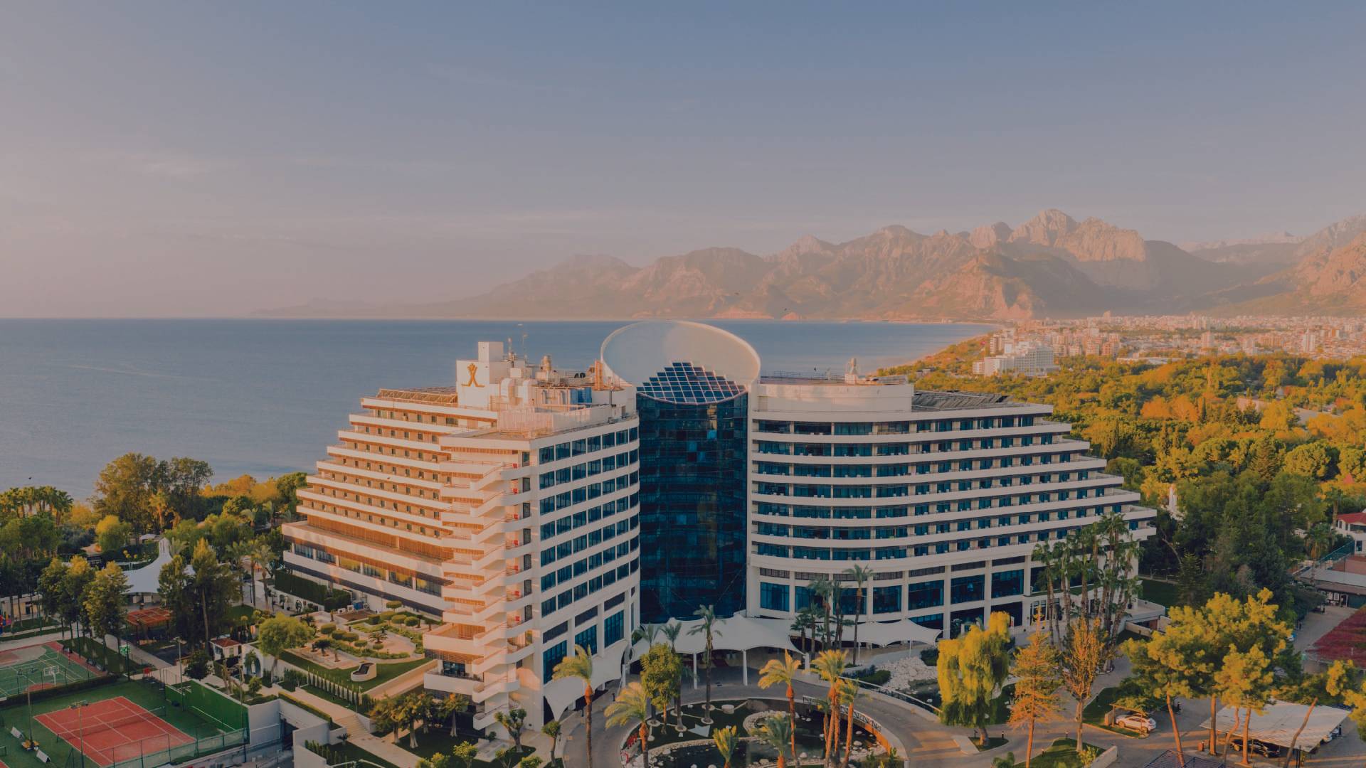 Aerial view of Rixos Downtown Antalya hotel with modern curved architecture, palm trees, and the Mediterranean Sea and Taurus Mountains in the background