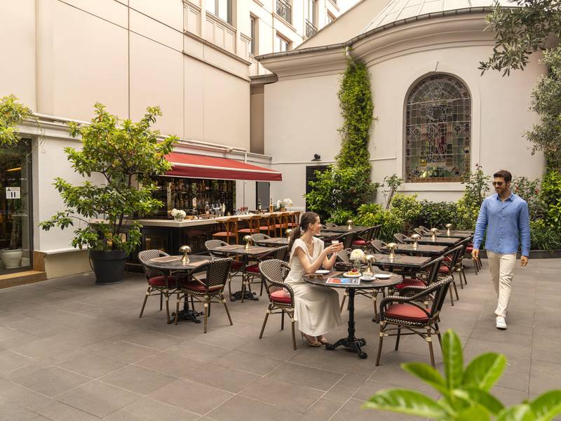 مقاعد في الفناء الخارجي لمطعم شابيل في مطعم ريكسوس بيرا إسطنبول، ويضم كراسي من الخوص، وباراً بمظلة حمراء، ومبنى تاريخي أبيض اللون مع نافذة كبيرة من الزجاج الملون. رجل يمر بجانب امرأة جالسة على طاولة.