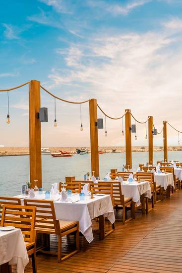 Elegant seaside dining setup at La Rosetta À La Carte Restaurant, Rixos Sungate, featuring wooden tables with white linens overlooking the marina and yachts under a bright blue sky.