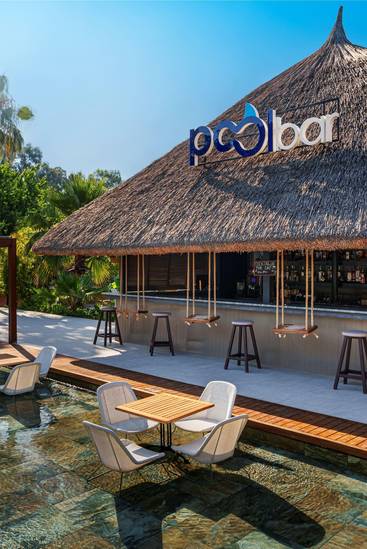 Thatched-roof Pool Bar at Rixos Göcek surrounded by lush greenery, featuring in-water seating, wooden decks, and hanging swing stools under a tropical-style canopy.