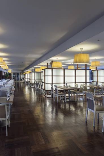 Rixos Sungate'teki Veranda Ana Restoran'ın iç görünümü, uzun ve modern bir yemek salonuna sahiptir. Koyu ahşap zemin, nötr tonlarda oturma grupları ve açık renk nevresimlerle döşenmiş masaların bulunduğu salon, bir sıra büyük, yuvarlak, bej gölgeli sarkıt lambayla aydınlatılıyor.